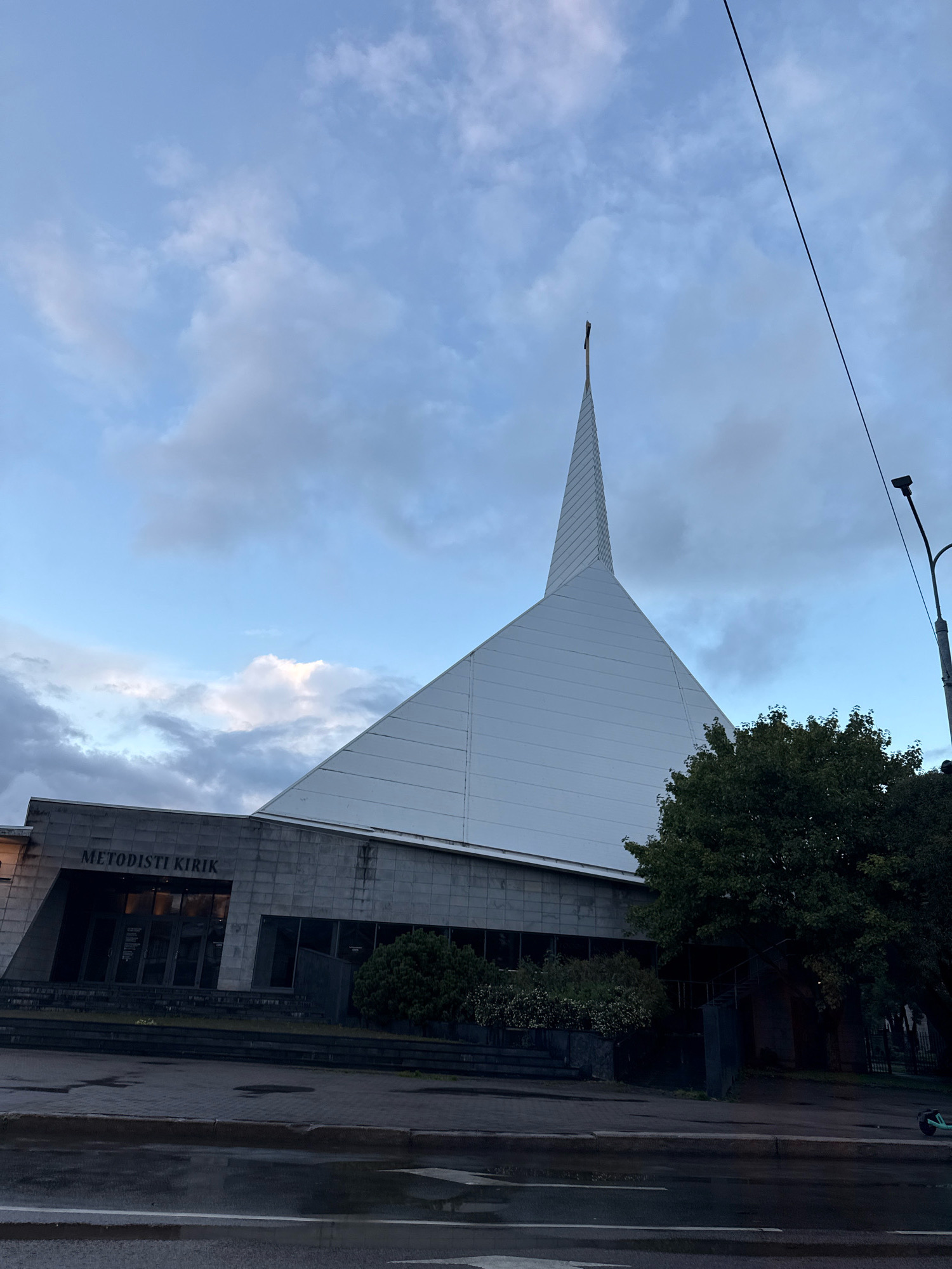 The image shows the Tallinn Methodist Church, located in Tallinn, Estonia. The church has a modern architectural design, characterized by a large, steeply pitched white roof topped with a cross. The facade of the lower section of the building is made of dark stone blocks. Above the entrance, the text "METODISTI KIRIK" is visible. The scene is set against a cloudy sky. In front of the church, there are steps leading up to the entrance, and some greenery, including a tree, is present on the right side of the building. The street in front of the church appears wet, suggesting recent rain. A street lamp is visible on the right side of the frame. There are no people or ongoing activities depicted in the image. The overall atmosphere is somber due to the overcast sky and wet conditions.