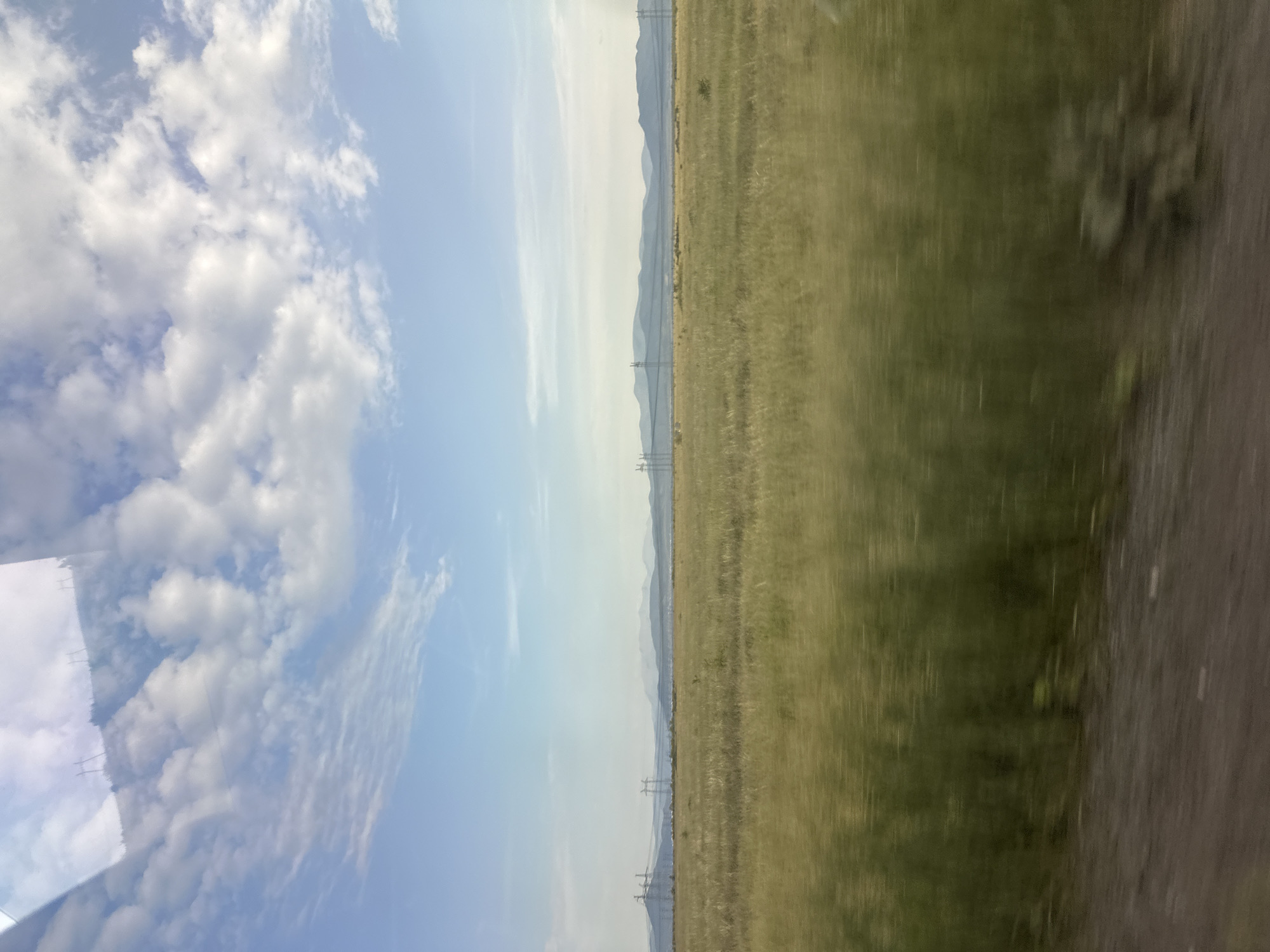 The image depicts a wide, open landscape under a sky filled with scattered clouds. In the foreground, a dirt road runs horizontally across the bottom of the frame. Beyond the road is a vast, grassy field, appearing somewhat blurred due to motion, suggesting the photograph was taken from a moving vehicle. The field stretches towards a distant horizon. In the mid-ground, several tall power transmission towers are visible, with power lines stretching between them. Behind these towers, a range of low, hazy mountains can be seen. The sky is a pale blue with white and light gray clouds. The overall scene is natural and rural, possibly a steppe or plains environment, and the presence of power lines suggests proximity to some form of infrastructure or development within a sparsely populated area. The light suggests it is daytime, likely late morning or afternoon. The image is taken from a slightly elevated perspective. There are no people or animals visible in the frame.