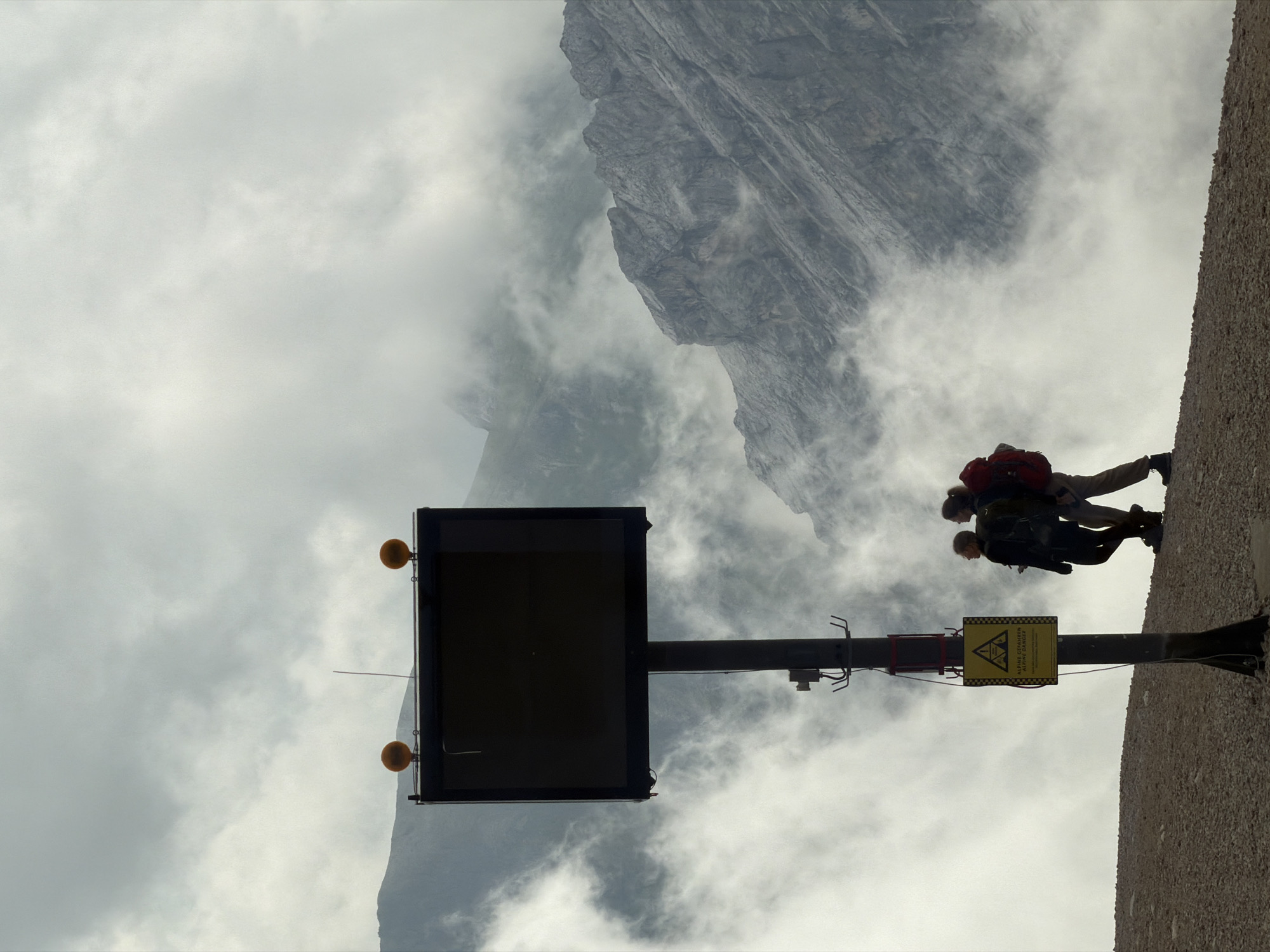 A silhouetted pair of hikers, one with a red backpack, stand near a sign and a large, dark electronic display board topped with two orange lights. The individuals are positioned on a rocky, elevated terrain, with a steep, mountainous landscape shrouded in fog and clouds in the background. The scene suggests a remote, high-altitude location, possibly a viewpoint or information point for trekkers in the Bavarian Alps near Garmisch-Partenkirchen, Germany, given the presence of the sign which reads "ALPINE GEFAHREN" (Alpine Dangers) in German. The overall atmosphere is somber and slightly dramatic due to the muted lighting and the encompassing fog.