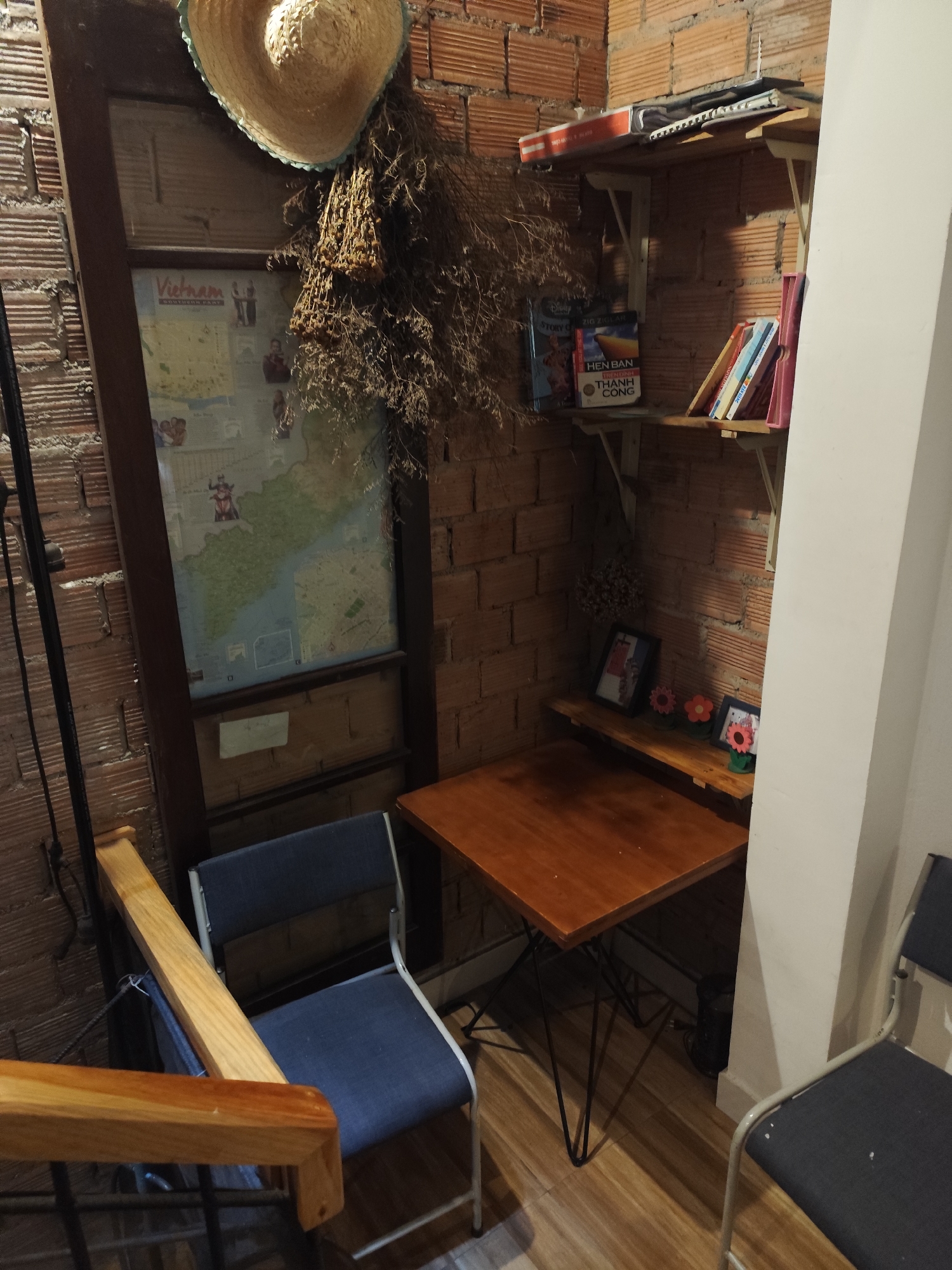 Indoor scene in Da Lat, Vietnam. Brick wall corner features a straw hat atop a framed map of Vietnam, accompanied by dried floral arrangements. Two wooden shelves hold books and picture frames. A small wooden table with hairpin legs sits between two chairs. One chair has a blue cushioned seat and a metal frame. The other chair has a dark gray seat and back with a metal frame.