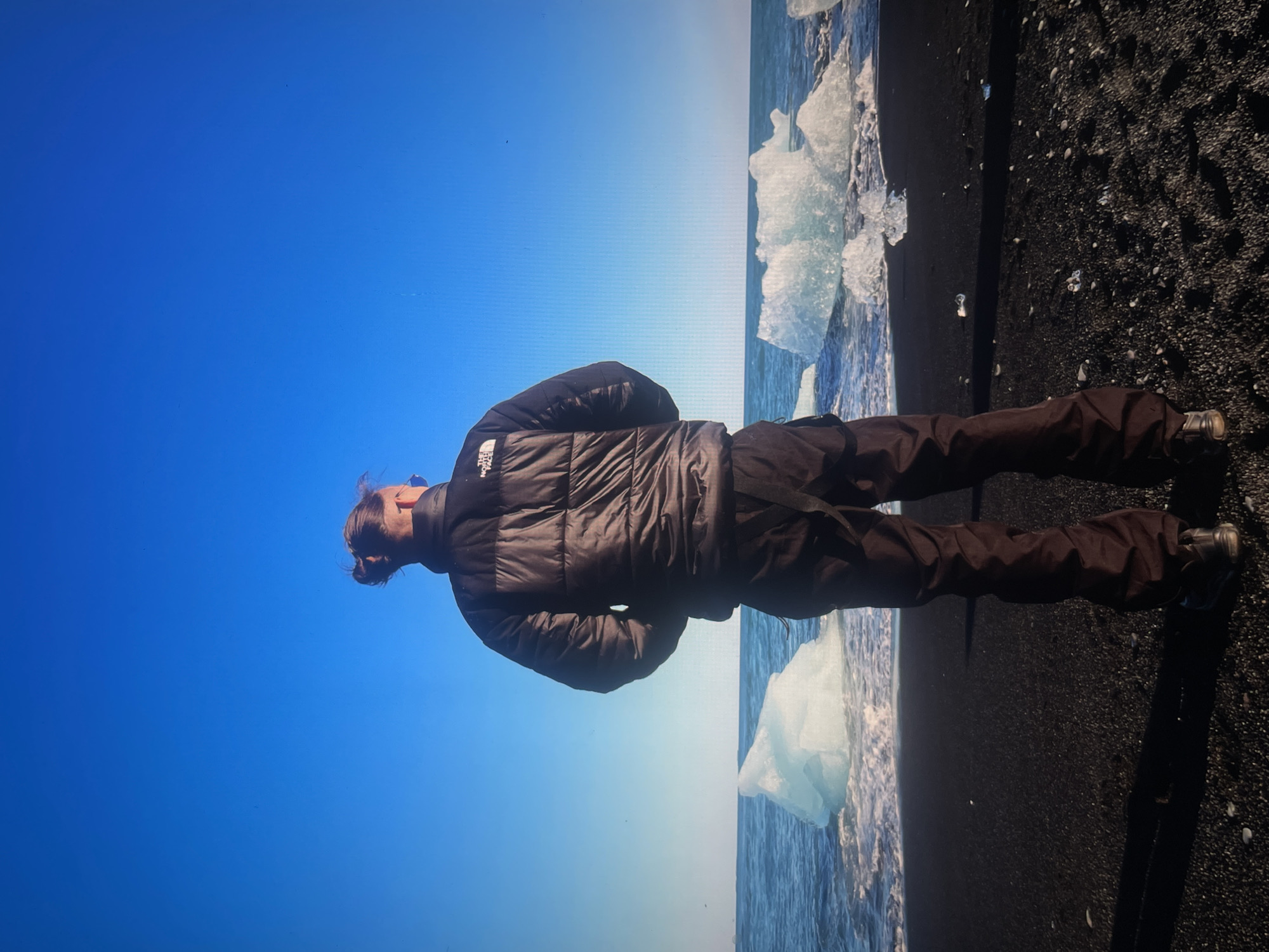 A person stands on a black sand beach next to ice fragments and ocean waves, beneath a clear blue sky. The person wears a dark jacket and dark pants. The location is not specified and is in the city of Unknown, Unknown.