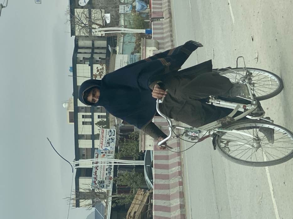 An individual is depicted riding a white bicycle on a paved road under an overcast sky in Kandahar, Afghanistan. The rider is positioned centrally, wearing a dark blue or black shawl that covers their head and most of their face, exposing only their eyes and forehead, consistent with male features. Beneath the shawl, they wear a brown long-sleeved garment and baggy brown trousers. Dark footwear is visible.
The bicycle has a white frame, silver handlebars, and a multi-bulb light fixture attached to the front. The individual's hands are gripping the handlebars, and their feet are on the pedals, indicating active propulsion.
To the left of the rider, a red and white striped concrete barrier separates the road from a sidewalk or shoulder. Beyond the barrier, multiple urban structures are visible. A prominent three-story building with horizontal window bands stands in the mid-background. Two large commercial billboards are present: one on the left advertises household appliances (partially legible text in Dari/Pashto includes "cooking utensils" and "complete goods"), and another on the right mentions "economy decoration." Bare trees, utility poles, and distant mountains are also visible under the muted light of the cloudy sky.