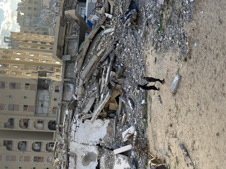 The image displays a scene of significant urban destruction in Khan Yunis, Palestinian Territory. In the foreground, two individuals are walking from left to right across a sandy, debris-strewn ground. The person on the left wears dark clothing; the person on the right wears a grey hooded sweatshirt and dark pants. Scattered on the sandy ground are small pieces of concrete debris, including a rectangular slab with exposed rebar. Immediately behind the individuals, the ground rises into a large expanse of collapsed building materials, consisting of concrete slabs, broken walls, and twisted rebar. This extensive rubble forms a significant barrier, obscuring the lower portions of structures in the background. Beyond the debris, multiple multi-story buildings exhibit severe damage, including large sections of missing walls, exposed interiors, shattered windows, and visible impact points on their facades. One prominent building in the background appears structurally compromised, showing a noticeable lean or partial collapse. The environment indicates extensive architectural devastation. The sky is partly cloudy, with sunlight illuminating the scene from the right.