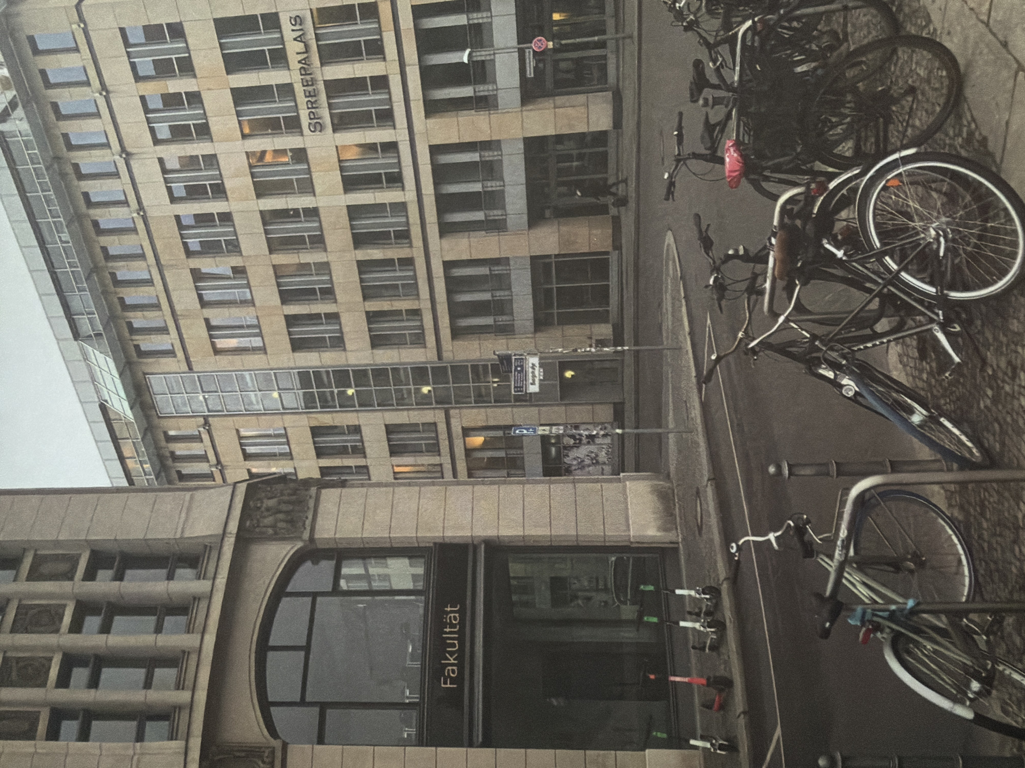 The image depicts a street scene in front of modern and classical buildings. On the left, a stone building with arched windows and decorative carvings is visible. A sign above an entrance reads "Fakultät." In the background, a taller building with a glass facade and a sign that says "SPREEPALAIS" dominates the scene. Several bicycles are parked on the sidewalk in the foreground. There are also a few electric scooters lined up near the entrance of the "Fakultät" building. The environment appears to be urban, with a street running in front of the buildings. No individuals are clearly visible or engaged in any distinct actions, besides one person walking in the background near the "Spree Palais" entrance. The lighting suggests it might be late afternoon or early evening. The city of Kyiv, Ukraine is not depicted in this image.