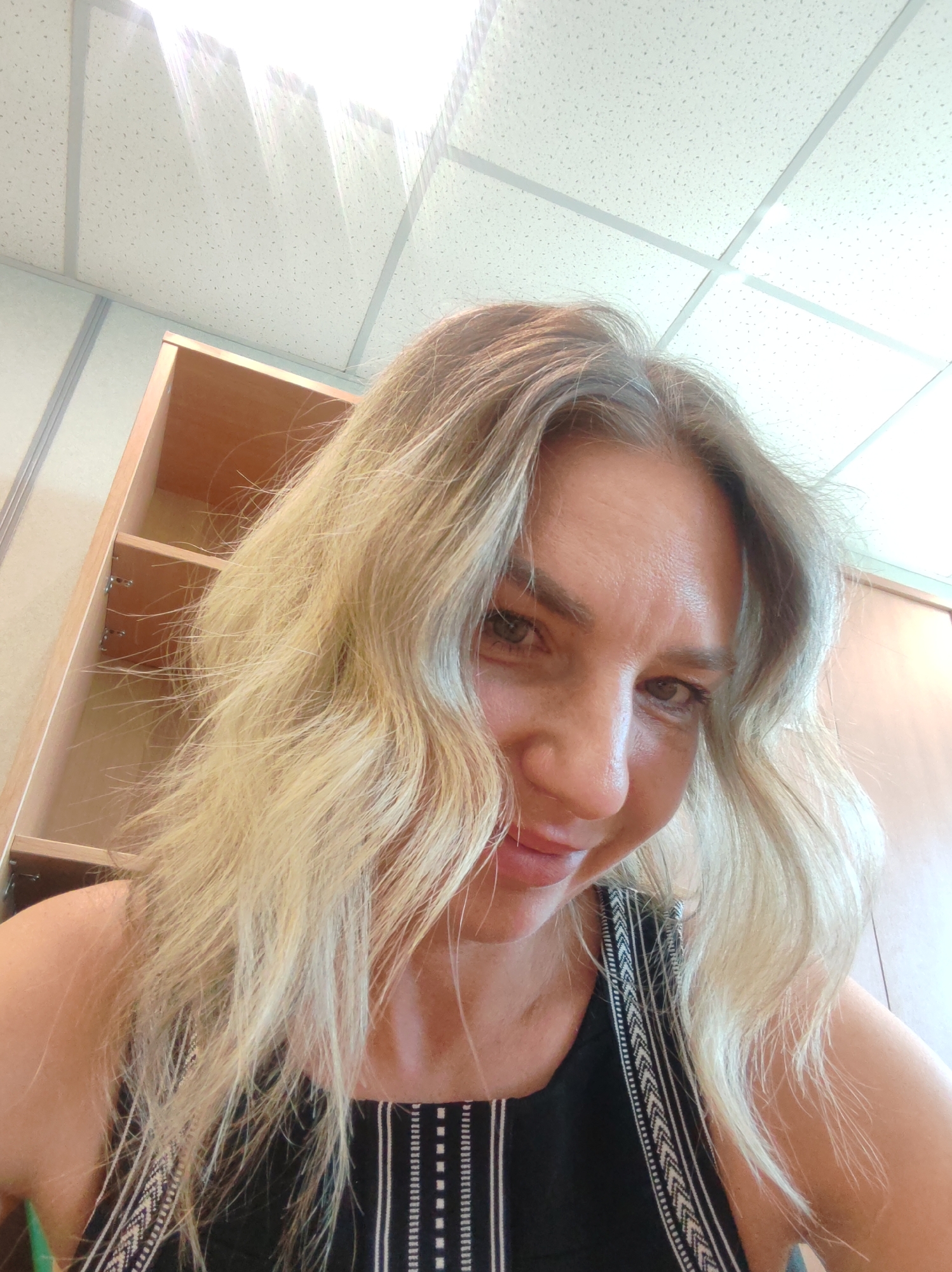 A light-skinned woman with blonde, wavy hair is seen in a close-up, selfie-style photo. She is wearing a black top with white vertical stripes and a geometric pattern along the edges. The background includes a ceiling with a grid pattern and fluorescent lighting. To the left, there's a partially visible wooden cabinet with shelves. Based on the appearance of the environment and lack of identifiable landmarks, it is not possible to confirm if the location is in Petropavlovsk-Kamchatsky, Russia.