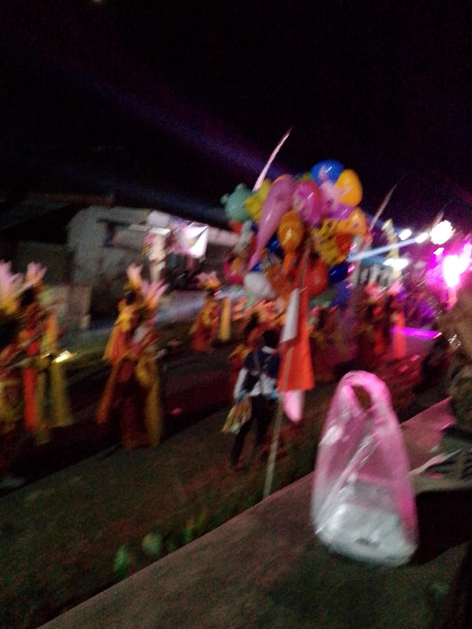 The image depicts a nighttime scene in Babadan, Indonesia, likely at a festive event. In the foreground, a transparent plastic bag containing white objects is visible on a dark, textured surface. The middle ground features several figures in what appear to be traditional costumes, adorned with colorful fabrics and headdresses, possibly engaged in a procession or performance. Behind them, a cluster of brightly colored balloons, including red, yellow, blue, and pink, is prominent. The background shows blurred structures, suggesting buildings or stalls, illuminated by artificial lights that cast purple and white beams across the scene. The overall impression is one of a lively outdoor gathering.