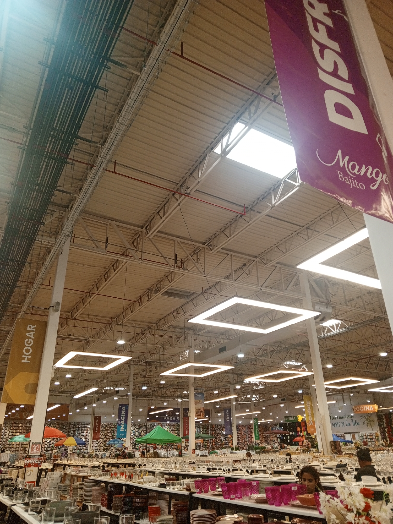The image shows the interior of a large retail store, possibly a department store or home goods store, in Pampatar, Venezuela. The high ceiling is exposed, revealing the building's structural elements like steel beams and ductwork. Square LED light fixtures are suspended from the ceiling, providing illumination. Merchandise is displayed on shelves and tables in the foreground, including glassware, plates, and other home goods. People are present in the scene, browsing the merchandise. Banners and signage hang from the ceiling, displaying the words "HOGAR", "REPOSTERÍA", "IMAGINA", "SONRIE", "COCINA", "FIESTA", "DINERÍA" and "DISFR".