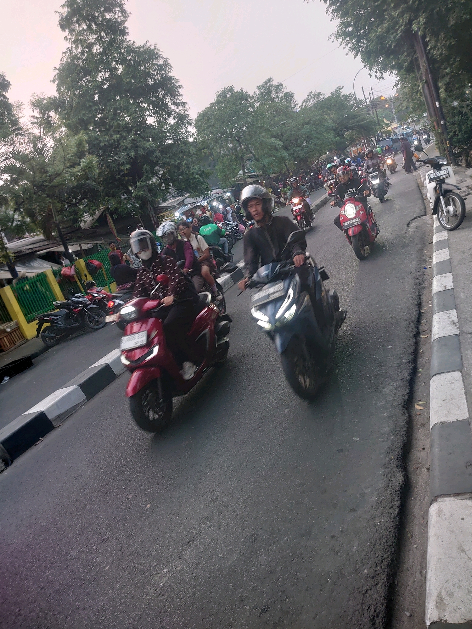 A group of motorcyclists, many wearing helmets, navigate a busy street in South Jakarta, Indonesia. The road is paved and appears to be a main thoroughfare, lined with trees on the left and a sidewalk with a curb on the right. Various motorcycles, including scooters and some with a more sporty design, are visible, some carrying multiple passengers. The scene captures the common sight of motorcycle traffic in urban Indonesia.