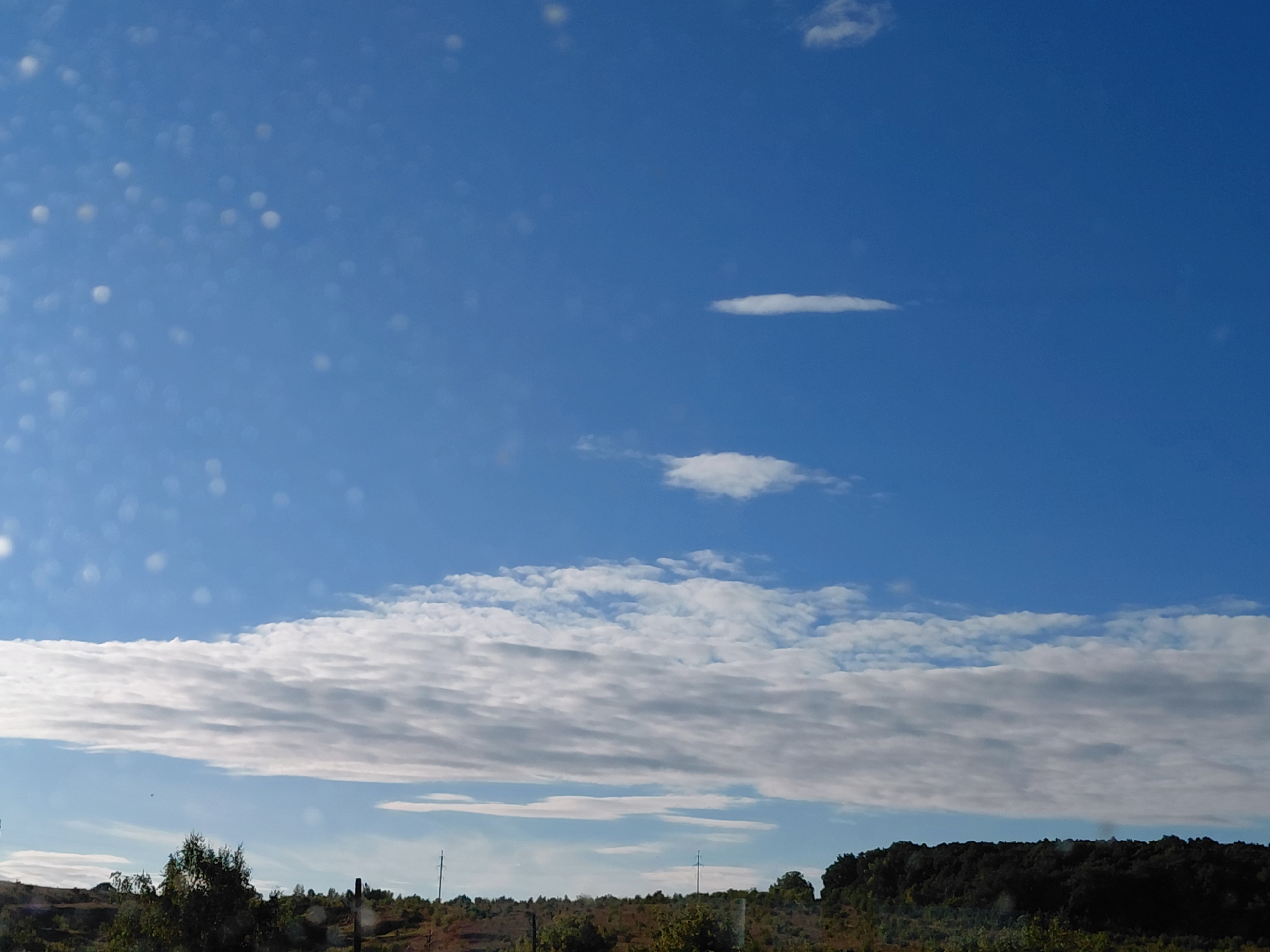The image shows a wide, unobstructed view of a bright blue sky with scattered clouds. A prominent, elongated cloud formation stretches horizontally across the middle ground, with lighter, wispier clouds above it. In the foreground, a varied landscape of rolling hills and vegetation is visible, including trees and bushes. Several utility poles are also present in the distance. The overall impression is a serene, natural landscape under a clear sky.