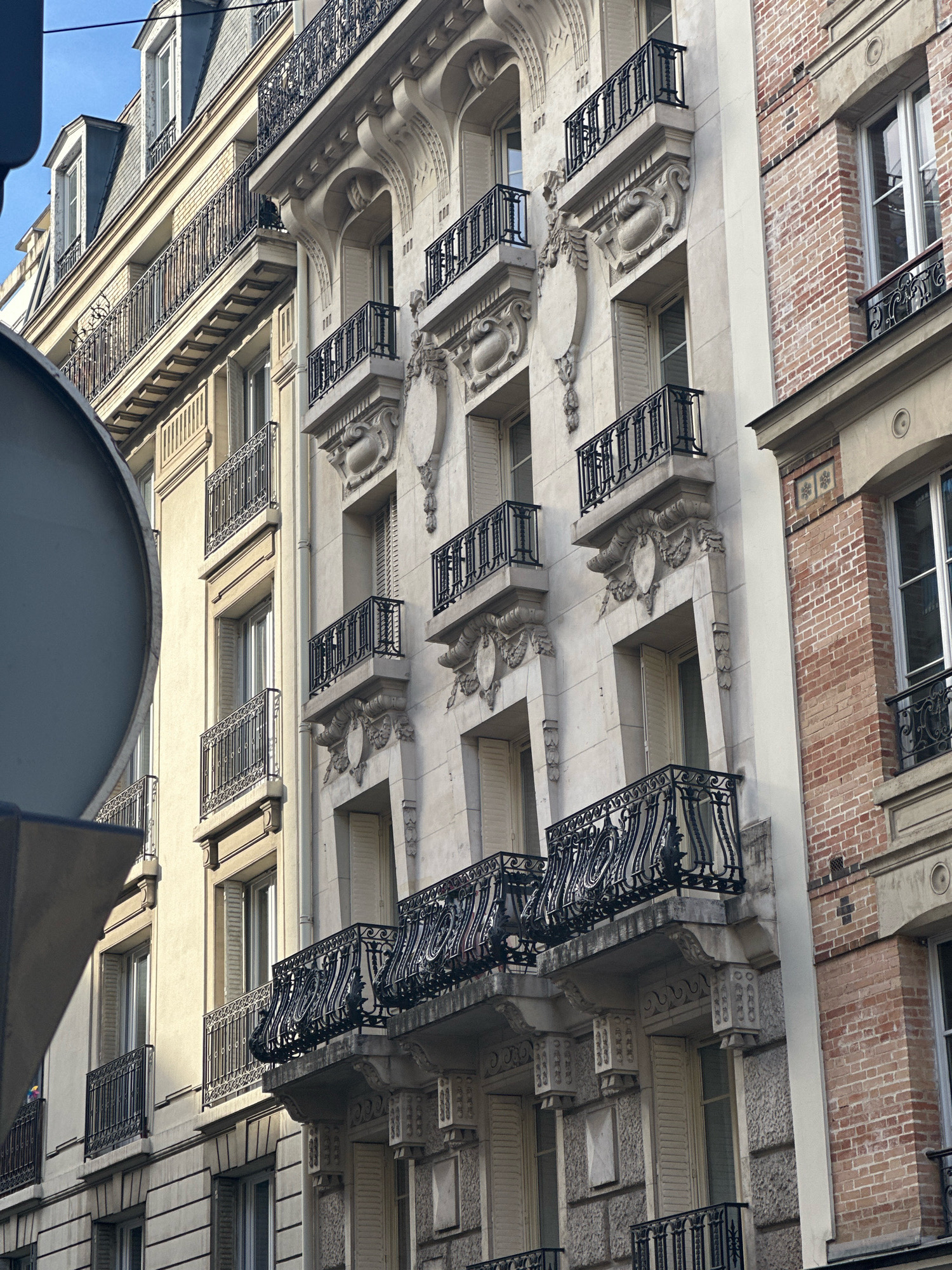 This image depicts the ornate facades of Haussmannian buildings in Paris, France, likely in a residential district. The architecture is characterized by a pale stone facade with classical decorative elements, such as sculpted reliefs above windows and doors, and wrought-iron balconies with intricate scrollwork. Multiple stories of windows, many with closed shutters, are visible. The building on the right contrasts with a facade of red brick. The sky above is a clear blue. The perspective suggests the photo was taken from street level, looking upwards at the buildings. A large, dark, rounded object, possibly part of a vehicle or street furniture, partially obscures the left side of the image. No people or specific activities are discernible.