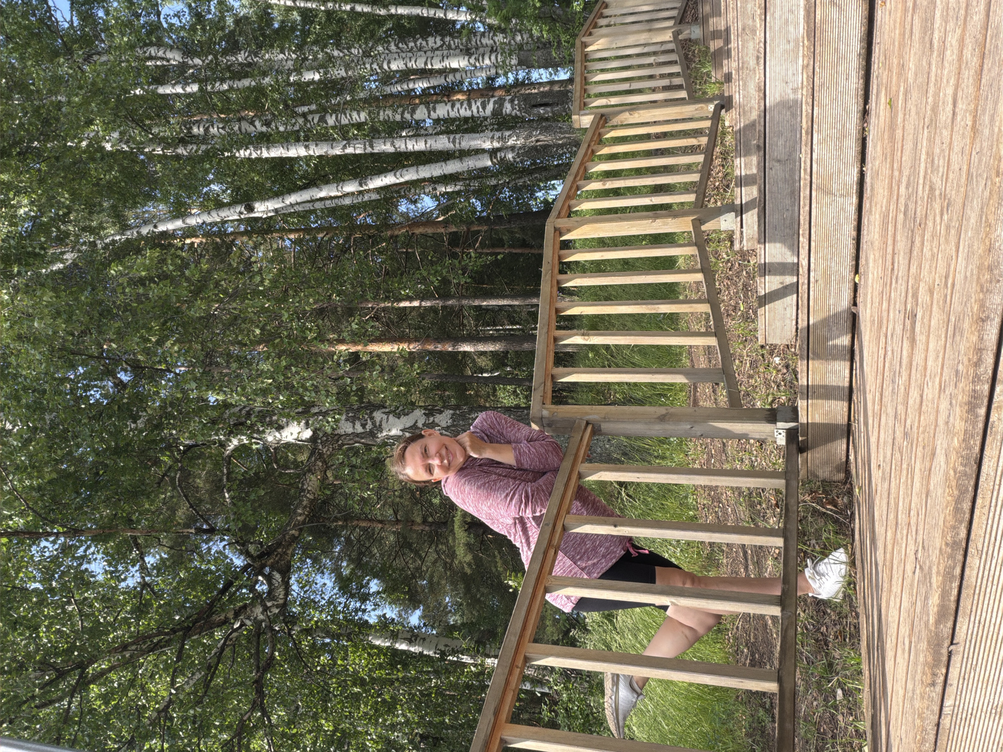 A woman is posing for a photo outdoors. She is leaning on a wooden railing, with her right leg bent and extended backward. She is wearing a pink and white marled hoodie, black shorts, and white sneakers. She has her chin resting on her hand and is smiling at the camera. The background consists of a wooden staircase and a dense forest with tall birch trees. The scene is set in a park or natural area in Chaikovsky, Russia. The lighting suggests it is daytime, likely with bright sunlight.