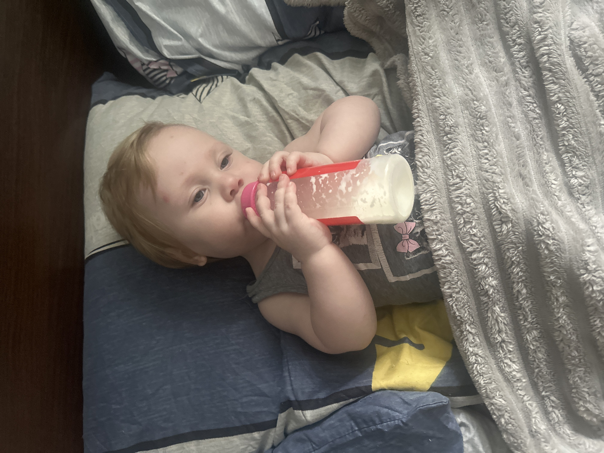 A toddler with blonde hair and a gray tank top is lying on a bed, drinking from a baby bottle. The bed has blue, gray, and yellow striped bedding, and a fuzzy gray blanket is covering the toddler's lower body. The toddler has a small red mark on their forehead. The image appears to be taken indoors, in a bedroom. There is no information in the image to suggest a location in Sayanogorsk, Russia.