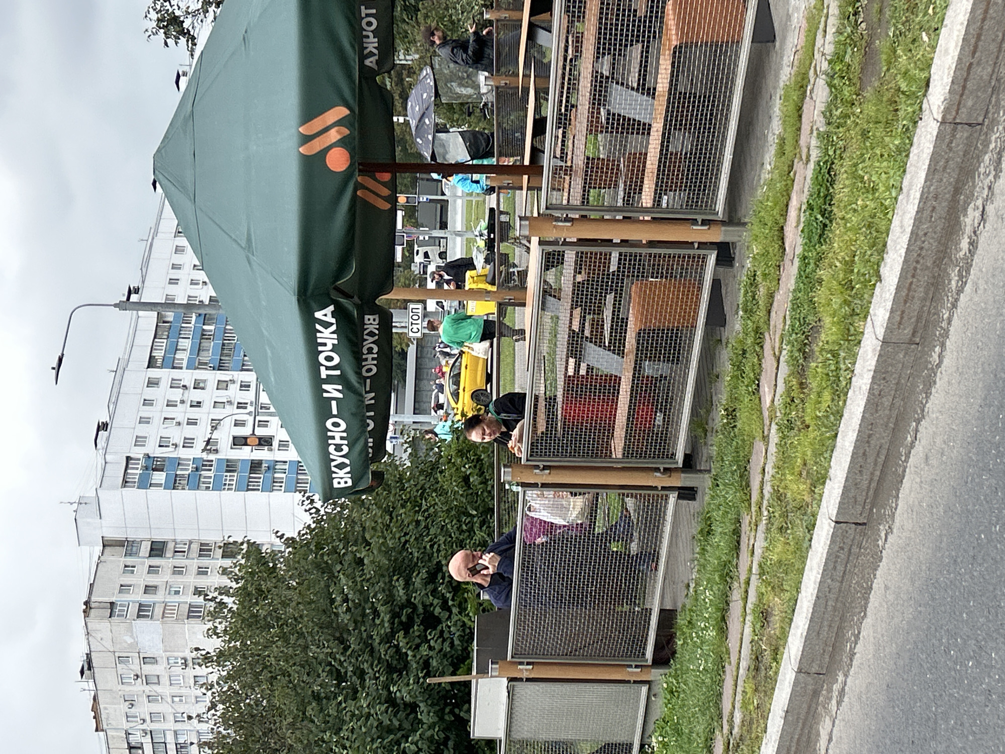 The image shows an outdoor seating area for a fast-food restaurant, likely a McDonald's given the iconic golden arches logo on the green awning, which also displays Cyrillic text reading "ВКУСНО — И ТОЧКА" ("Tasty – And Point"). The awning is large and covers several picnic tables. In the foreground, a gray concrete curb runs along a street, with a grassy area and a paved walkway behind it. A metal fence with mesh panels encloses the seating area.
On the left, a bald man with a dark shirt is sitting at a picnic table and talking on a mobile phone. He has a plastic bag on the table. To his right, a woman in a black hoodie is also seated and appears to be eating. Behind them, a bright yellow car is visible on a street, suggesting the location is in a urban or suburban area. In the background, several multi-story apartment buildings are prominent, some with white and blue facades. The sky is overcast.
Further to the right, other patrons are visible, including a person under a black and white umbrella, and another individual in a blue jacket. Traffic and more buildings are discernible in the background. The overall scene depicts a casual dining environment in Moscow, Russia, likely near a residential area, on a cloudy day.