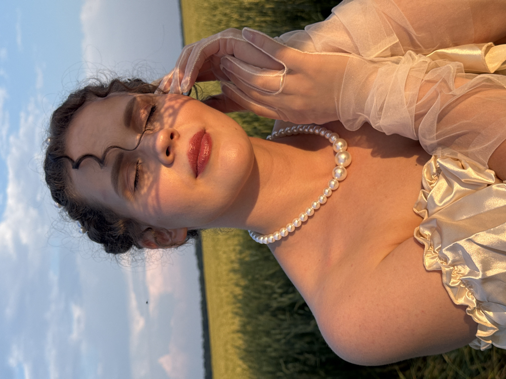 A young woman with curly dark hair styled in braids and adorned with small pearls is positioned in the foreground, facing slightly to the left with her eyes closed. She wears a pearl necklace and sheer, ruffled white gloves. A shadow of leaves is cast across her face and décolletage. She is dressed in an off-the-shoulder satin garment with ruffled trim. The background features a field of golden wheat under a cloudy sky, bathed in the warm light of sunset. The scene appears to be a portrait taken outdoors in a rural setting, possibly Malyi Bukor, Russia.