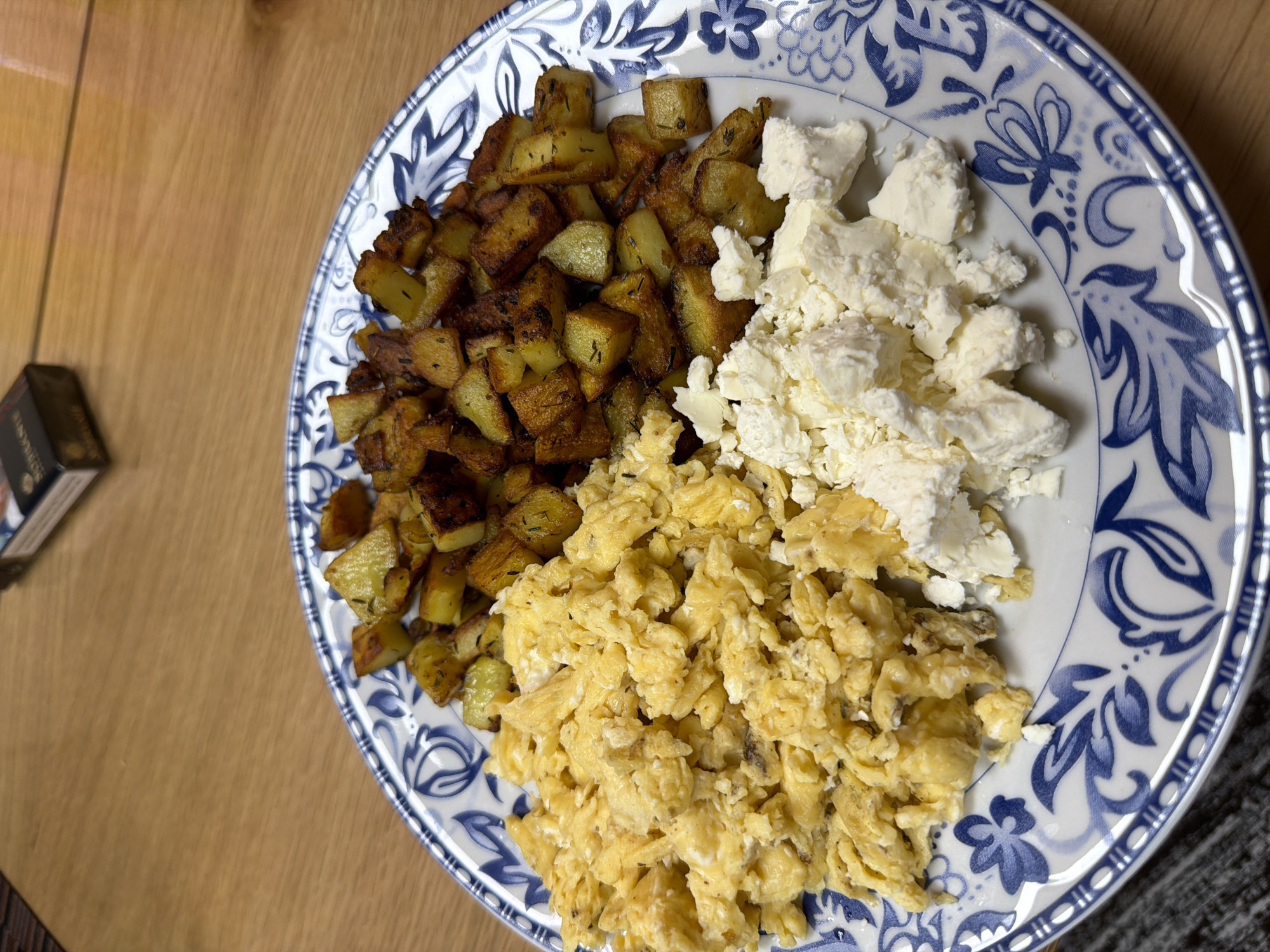 On a wooden surface, a plate with a blue and white floral pattern holds three distinct food items. To the left, diced and seasoned potatoes are visible, appearing golden brown and slightly crispy. Adjacent to the potatoes, crumbled white cheese is situated, its texture appearing soft and moist. Occupying the lower portion of the plate are scrambled eggs, their yellow color suggesting they have been properly cooked. To the left of the plate, a small, dark-colored box is situated on the wooden surface. The scene lacks context to determine the specific location in Panagyurishte, Bulgaria, or any other detailed information.
