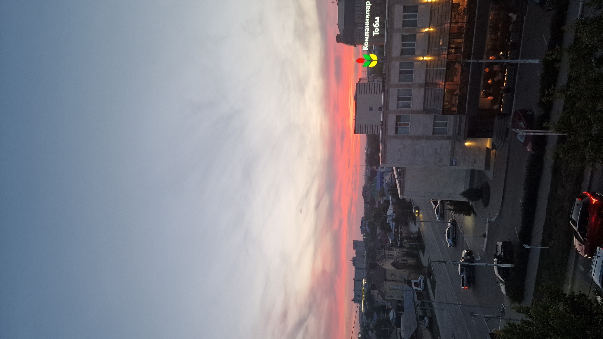 This aerial photograph captures a cityscape at dusk in Kostanay, Kazakhstan. The sky is a gradient of pale blue to soft pink and orange hues, indicating the setting sun. The foreground features a multi-story building with a glass-enclosed restaurant or establishment on the ground floor, illuminated by warm interior lights. Above this building, a sign displays a logo with a red and green plant-like symbol and text in Cyrillic which reads "Компаниялар Тобы".
To the left, a street is visible with several cars. A red SUV is parked in the lower center, its brake lights illuminated. Other vehicles, including a pickup truck and sedans, are seen on the street and at an intersection. Beyond the street, residential houses with pitched roofs and some larger industrial-looking buildings are scattered across the landscape. Trees and greenery are present along the street and in the distance. The overall scene depicts a city transitioning from daylight to evening, with artificial lights beginning to illuminate the buildings and streets.