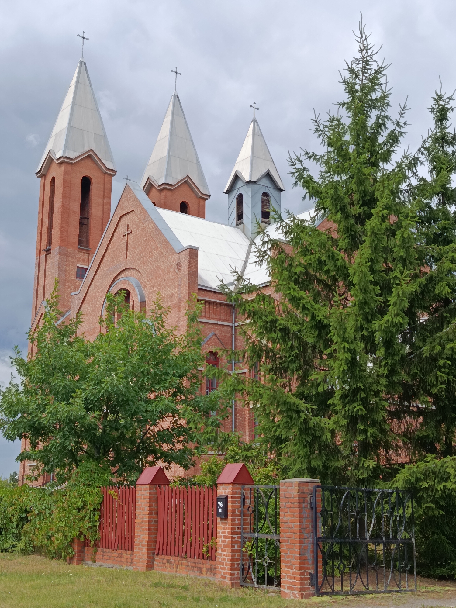 The image shows a brick church with a complex roofline and multiple spires, some topped with crosses. The church is partially obscured by lush green trees, including a prominent evergreen on the right side. In the foreground, a red wooden fence with brick pillars runs horizontally across the frame. One pillar has a black mailbox with the number "76" and a letter "a". The sky above is overcast with gray clouds. The scene appears to be outdoors on a cloudy day. This is likely the Catholic Church of the Holy Ascension in Svietlahorsk, Belarus, given the architectural style and common features of churches in the region.