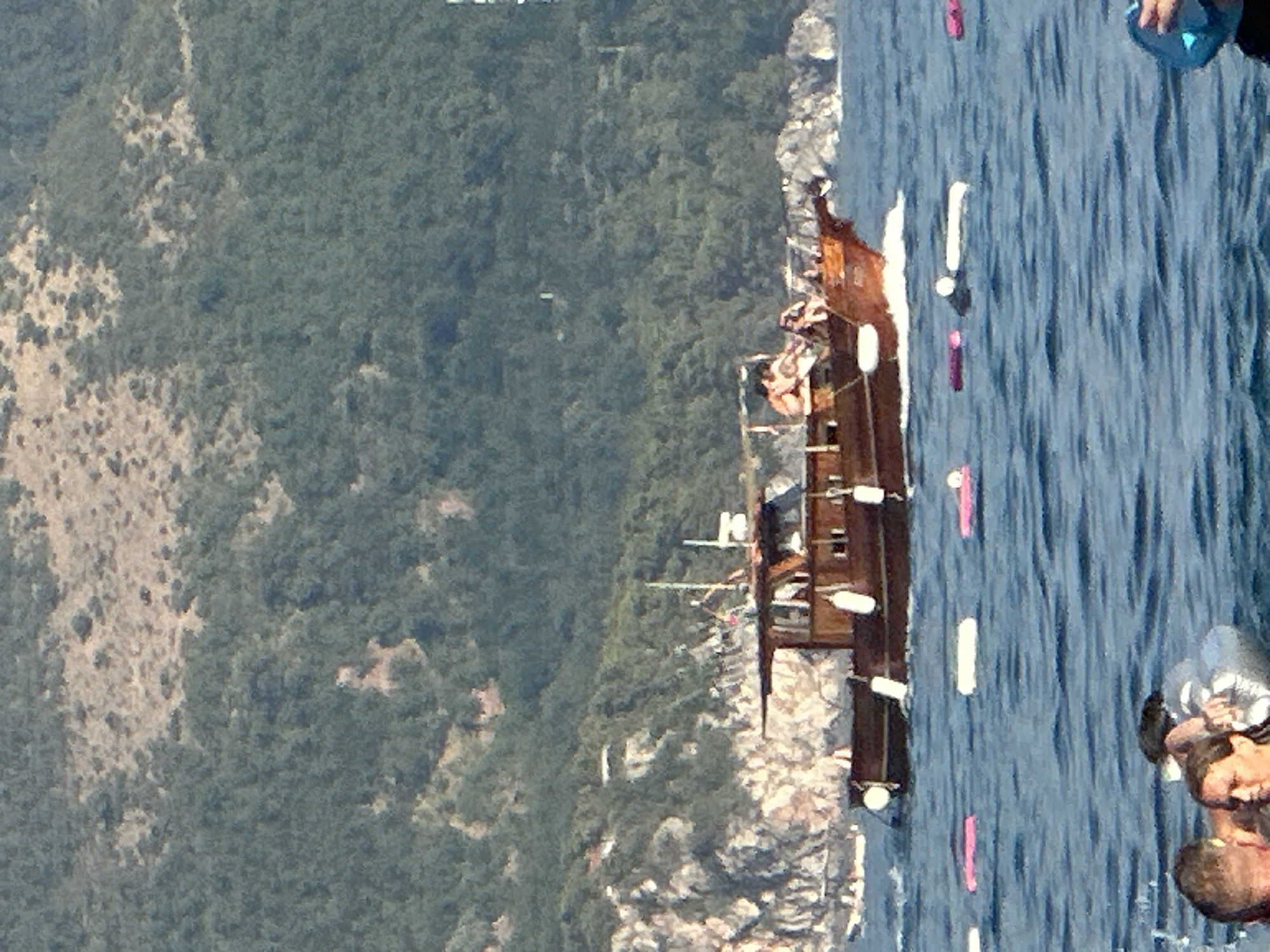 In the waters off the coast of Boreti, Montenegro, a wooden boat with several passengers is seen cruising near a rocky, tree-covered shoreline. The boat appears to be a tour or pleasure craft, with people relaxing on its deck. In the foreground, several inflatable buoys, some pink and some white, are floating on the water. The water is blue and choppy, indicating a slight swell. The overall scene depicts a leisurely day at sea, with the natural beauty of the Montenegrin coast as a backdrop.