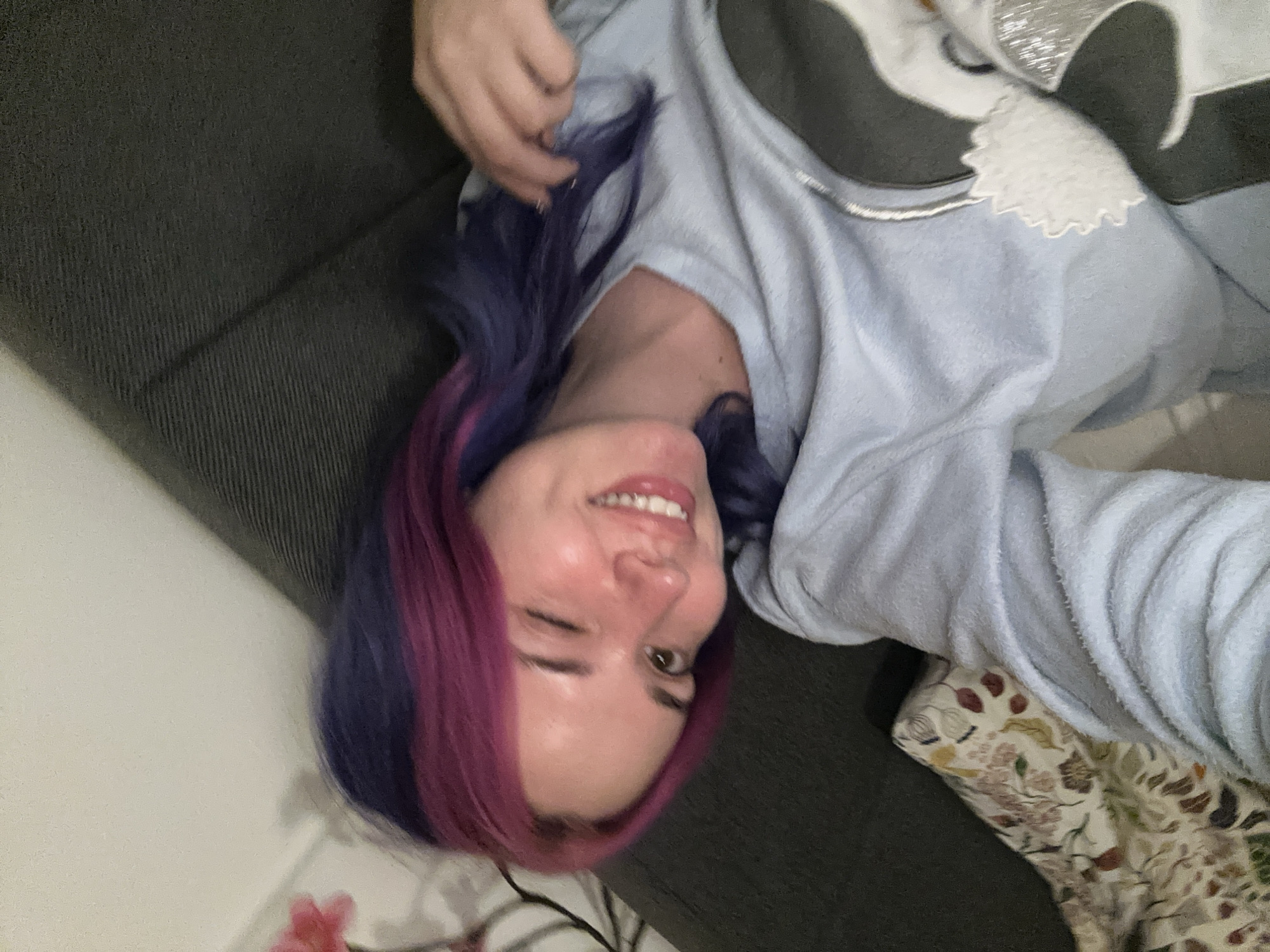 The image captures a young woman in an indoor setting, likely taking a selfie. She is positioned on a dark grey sofa, with the camera held from an overhead, slightly diagonal angle. The woman has distinctive two-toned hair, a mix of vibrant purple and magenta or pink, which falls around her face. She is wearing a light blue, soft-textured top, possibly made of fleece or similar loungewear fabric, featuring a white and grey design on the shoulder area that appears to be a stylized animal, possibly a wolf or dog, with some shiny embellishments.
Her expression is joyful and playful; she is smiling broadly with visible teeth and winking with her right eye. Her left hand is raised near her head, gently touching her hair. Next to her, a patterned pillow or blanket with a floral or leafy design in muted colors (beige, brown, green, red) is visible. A plain light-colored wall forms part of the background. The lighting suggests an indoor environment, possibly under artificial light. No visible text is present in the image. The overall mood conveyed is relaxed and cheerful.