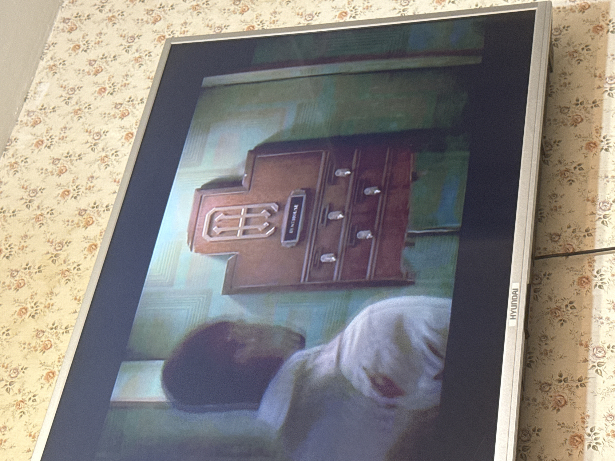 A silver-framed Hyundai television is displaying a scene from a movie or television show. The scene features a brown, wooden device labeled "Guesthouse" mounted on a wall. Below the device, a person is sleeping in a bed. The television is positioned in front of a wall covered in floral wallpaper. The location cannot be determined from this image.