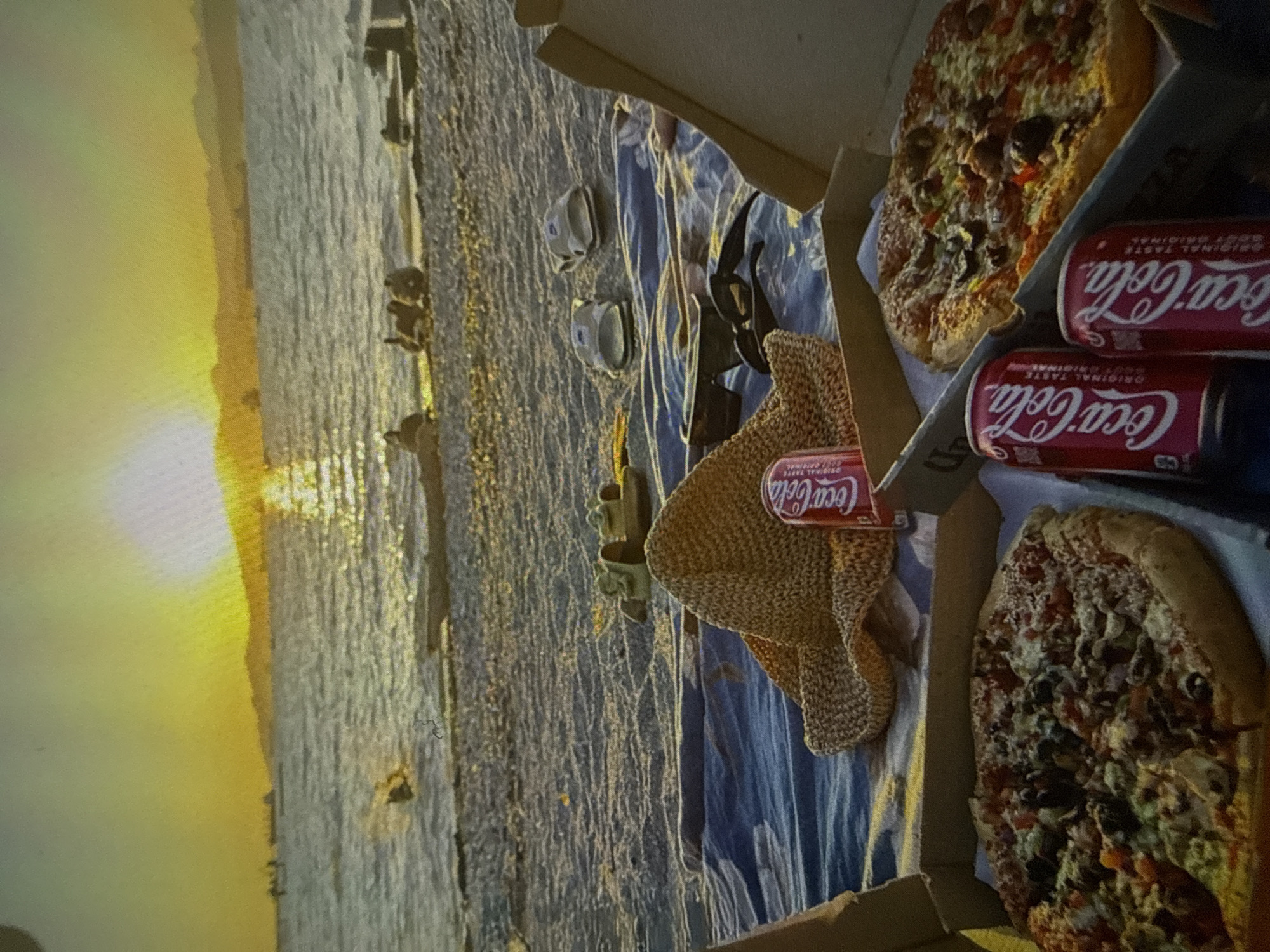 This media file captures a serene sunset scene on a beach, likely in Varna, Bulgaria, showing a relaxed outdoor picnic setup. The foreground features a blue patterned blanket or towel laid out, holding several items for a meal.
Key subjects include two large pizzas, partially visible in cardboard boxes, suggesting they are take-out. Alongside the pizzas are three cans of Coca-Cola, two placed upright next to one pizza box, and a third resting on a straw hat. The text "Coca-Cola" and "ORIGINAL TASTE" is clearly visible on the cans. Other objects on the blanket include a pair of dark sunglasses and two small, white containers or cups.
The activity taking place is a casual outdoor meal or picnic, set against a picturesque backdrop. The time of day is clearly sunset, as indicated by the brilliant golden and orange light spread across the horizon, casting warm reflections on the vast body of water, which appears calm. The sky is bright and clear, enhancing the tranquil atmosphere. There are no people visible in the frame, allowing the focus to remain on the meal and the beautiful natural setting. Distant structures or landmasses are faintly visible on the horizon, outlined by the setting sun.