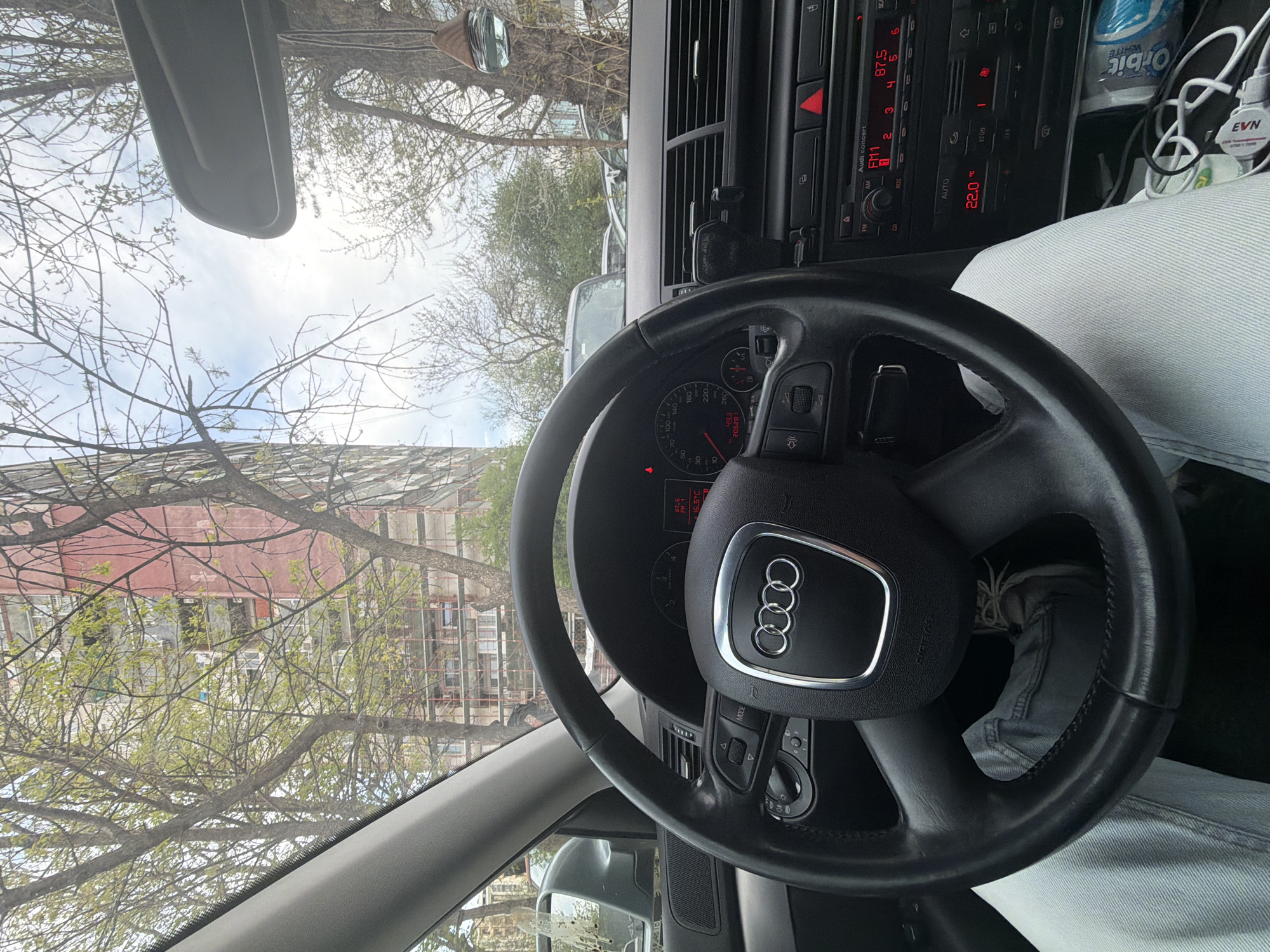 The image is taken from inside a parked Audi car, focusing on the steering wheel and dashboard. The steering wheel is black with the Audi logo in the center. The dashboard features a car stereo displaying radio information, with the frequency 87.5 FM1 selected, and the temperature set to 22.0 degrees Celsius. A pack of Orbit White chewing gum and an EVN-branded charger are visible on the dashboard next to the radio. The driver is wearing light blue denim pants. Outside the car window, there are trees with budding leaves and a building in the background, suggesting an urban environment. Based on the architecture and environment, the location is identified as Plovdiv, Bulgaria.