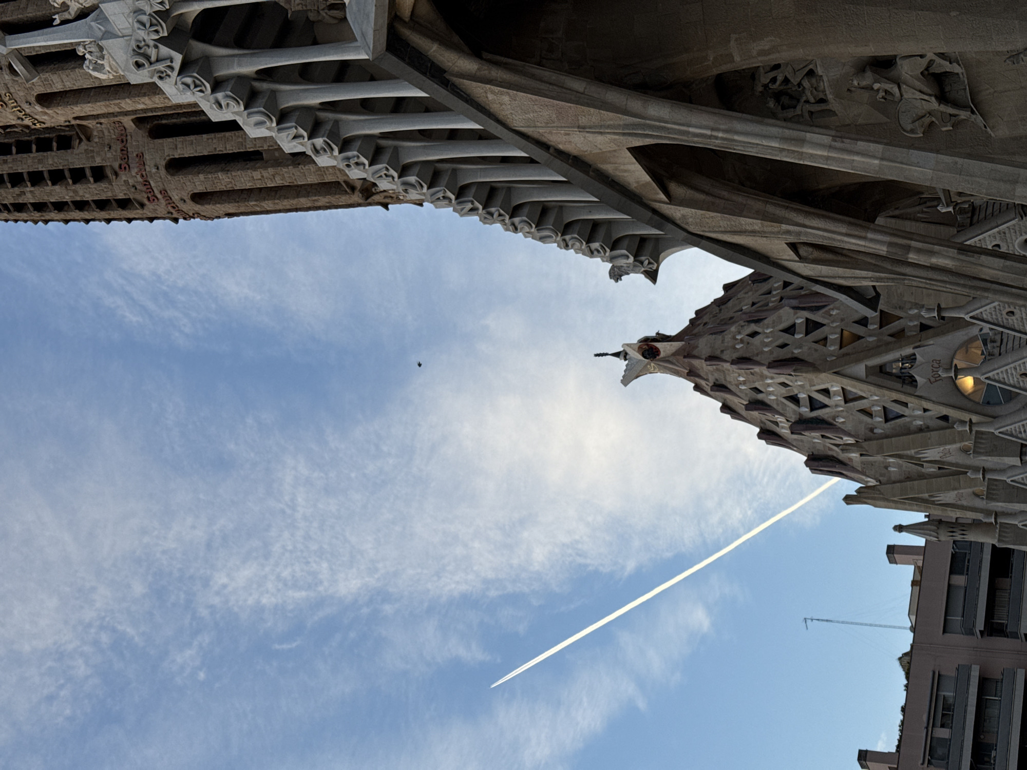 An upward-angled view captures elements of the Sagrada Família basilica in Barcelona, Spain. The primary subjects are a portion of the Passion Façade and one of its bell towers, set against a blue sky with scattered cirrus clouds.
The bell tower, located on the far right, is constructed of reddish-brown stone with vertical rectangular apertures. The word "Sanctus" is inscribed multiple times in red text on its surface. Adjacent to the tower, the Passion Façade displays a distinct angular, geometric stonework pattern, featuring multiple sculpted figures, arches, and buttresses. A spire-like element on a lower section of this façade shows a red or orange accent at its peak, and a cross finial is visible above another section of the facade.
The sky is clear blue with wispy white clouds. A straight, white aircraft contrail extends diagonally from the lower-left toward the upper-right. A small, dark bird is present in the upper-left sky. In the lower-left background, parts of modern urban buildings with balconies and flat roofs are visible. A slender, tall antenna rises from one of these rooftops, accompanied by several chimneys.