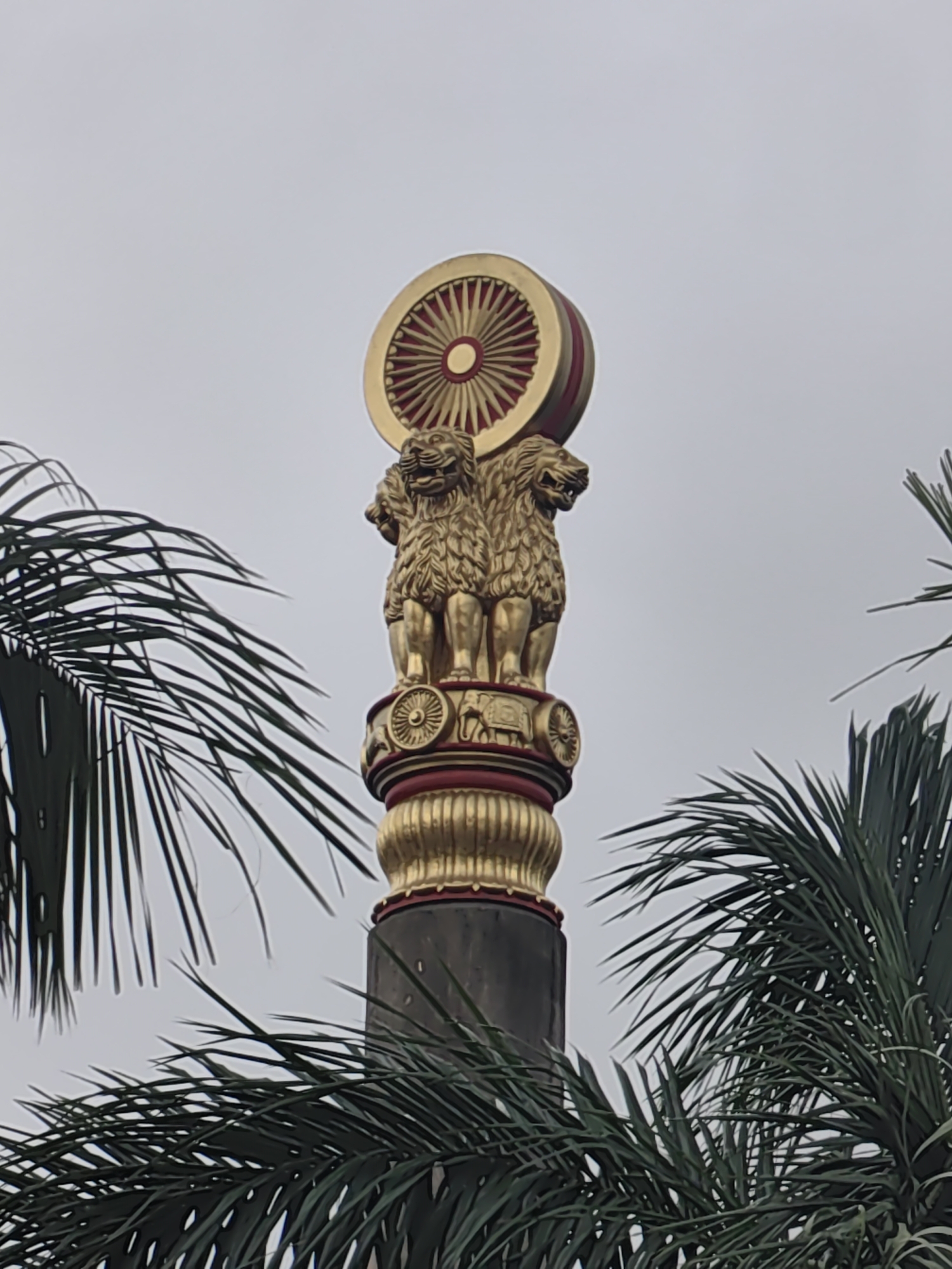 The image shows a close-up of the Ashoka Pillar, a significant national symbol of India, set against a cloudy sky. The pillar, depicted in gold and red, features four lions at the top, facing in different directions, with a Dharma Chakra (wheel of law) above them. The base of the pillar has decorative bands and animal carvings. In the foreground and partially obscuring the pillar on both sides are the fronds of palm trees, suggesting an outdoor setting. While the image is cropped, it is highly likely that this is a depiction of the Ashoka Pillar located at the Gateway of India in Mumbai, India, a prominent landmark in the city. There are no people visible in the frame.