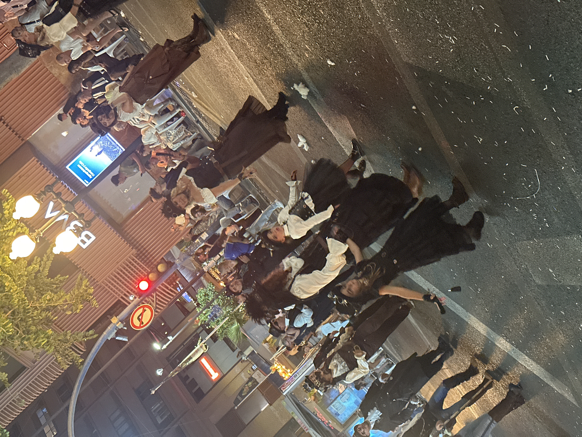 The image captures a nighttime street scene in Alicante, Spain, during a public event or parade. Numerous individuals are visible, with approximately a dozen in the foreground and midground dressed in elaborate, dark-toned costumes resembling Victorian or steampunk styles, featuring long coats, corsets, ruffled blouses, top hats, and boots. These costumed figures are actively moving along an asphalt street, which is strewn with scattered white, elongated confetti or paper debris. On the right side of the street, several spectators in casual modern attire are seated on white plastic chairs or standing on the sidewalk, observing the activity.
The urban environment includes multi-story buildings lining the street, one featuring a distinct ribbed architectural facade. A brightly illuminated digital display board on a building wall is visible, showing text in Spanish, including "Acapulco Capella y Calle," alongside an image. Street lighting from multiple lampposts illuminates the scene, casting shadows. A red traffic light is present on the left, along with a circular red traffic sign depicting a curved arrow, indicating a traffic restriction. Overhead, foliage from trees is visible against the night sky.