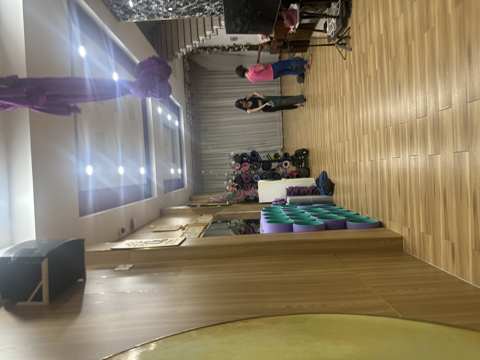 This image shows the interior of a building, possibly a yoga studio, located in Bac Nha Trang Ward, Vietnam. Two people stand in the center of the room, one wearing a pink shirt and blue pants and another wearing black pants and a black shirt, and appear to be conversing or interacting in some way. In the foreground, there is a series of purple yoga wheels with green centers arranged neatly on a raised platform. Further back, there is a rack filled with yoga mats and other fitness equipment. In the background, curtains hang along a wall, and light fixtures are seen on the ceiling. Additional furniture and objects are visible in the background. The floor is made of wooden planks. A purple aerial yoga hammock is hanging from the ceiling in the upper left. There are frames hanging on the wall and a black cushioned bench to the left of the image. The lighting in the studio is provided by ceiling-mounted fixtures.
