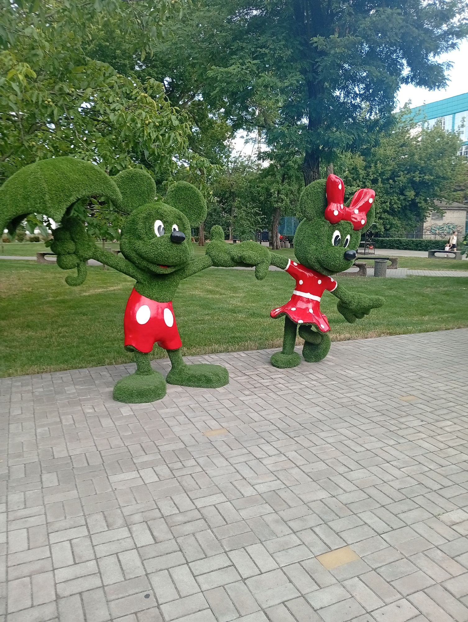 In the city of Pokrov, Ukraine, two topiary figures resembling Mickey Mouse and Minnie Mouse stand on a paved walkway. The figures are sculpted from green shrubbery, with the Mickey Mouse figure wearing red shorts with white polka dots and the Minnie Mouse figure wearing a red dress with white polka dots and a red bow with white polka dots in her hair. They are positioned in a park-like setting with green grass, trees, and benches visible in the background. A building with a blue facade is also visible in the distance. The paving stones are light gray and arranged in a geometric pattern. The overall scene depicts an outdoor decorative display in a public space.