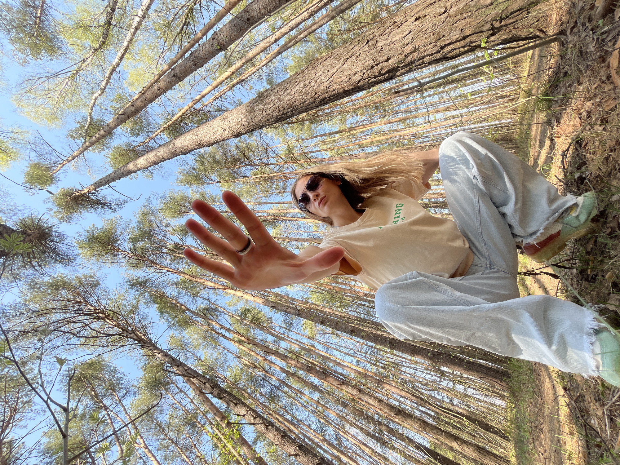 The image is a low-angle perspective photograph taken in a forest in Chaikovsky, Russia. A young woman is the primary subject. She is wearing sunglasses, a beige t-shirt with green lettering, light blue jeans, and green shoes with red soles. She has blonde hair. Her left hand is extended towards the camera, palm open. The background consists of tall, thin trees reaching towards a blue sky. The ground is covered with dry leaves and other natural forest debris.