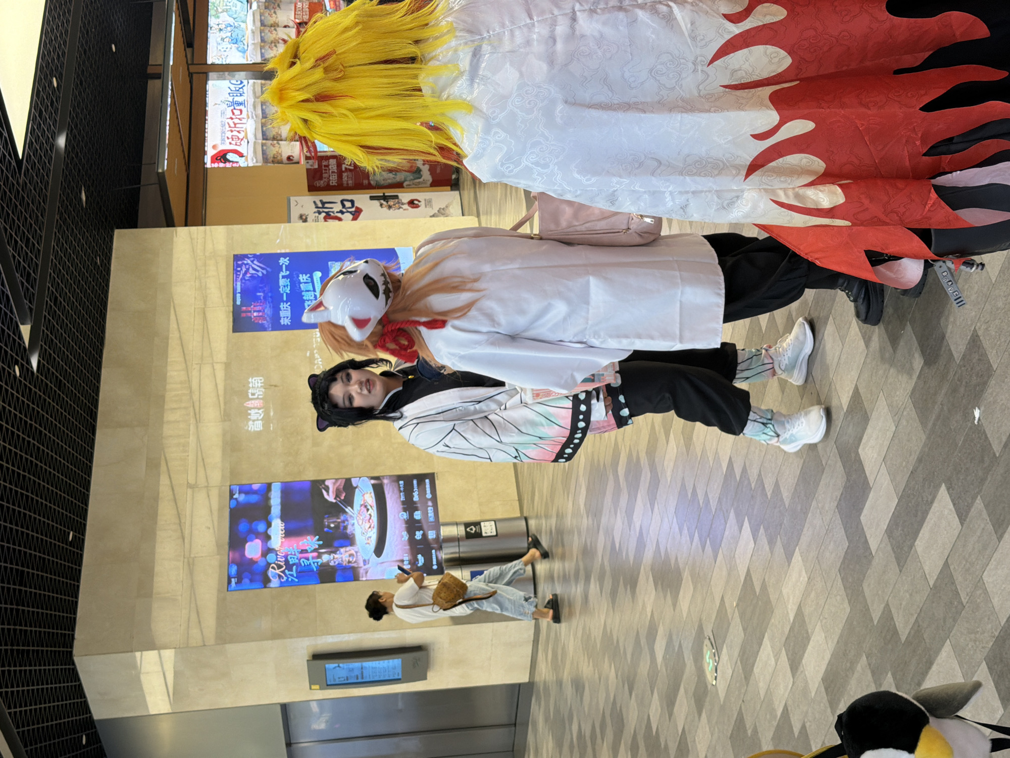 In Yuzhong District, China, three individuals are present in a brightly lit, indoor commercial space with a tiled floor. Two individuals are in the foreground, facing away from the camera, and are dressed in anime-inspired cosplay outfits. The person on the right has bright yellow hair and a white robe with a red flame pattern. The person in the middle has orange hair and wears a white robe with a fox mask and red accents. To the left, a third individual with black hair and cat ear accessories is facing the other two and appears to be engaged in conversation. In the background, a woman with her hair in a bun is walking away from the camera, carrying a small brown bag. Digital advertising screens are visible on the wall behind the cosplayers, displaying images and text. The environment suggests a public area, possibly a shopping mall or exhibition center.