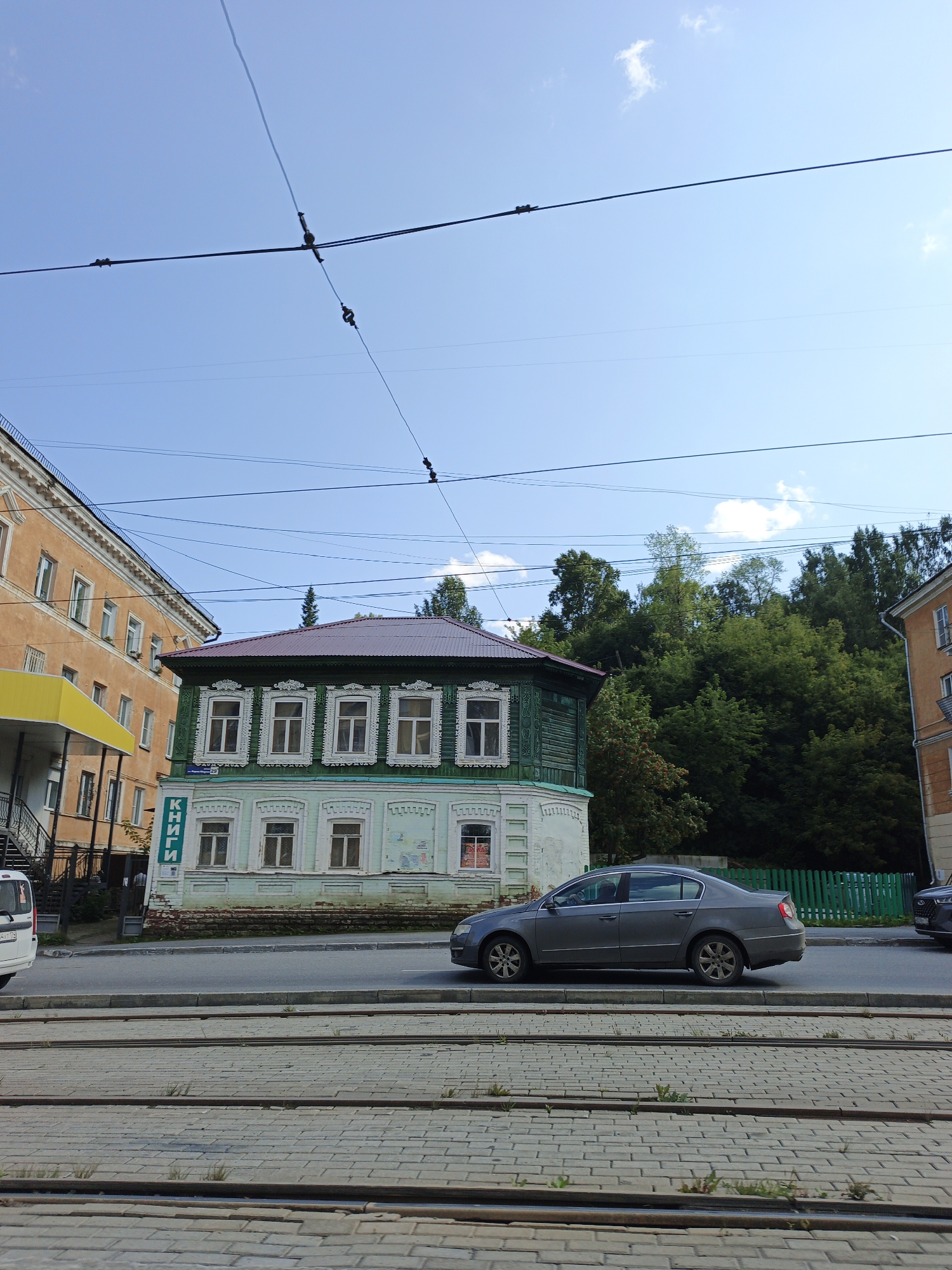 The image depicts a street scene in Zlatoust, Russia, on a bright, sunny day with a clear blue sky dotted with a few small clouds. The foreground is dominated by a paved street with visible tram tracks running horizontally across the frame. Beyond the tracks, a two-story building with green wooden siding on the upper floor and light blue plaster on the lower floor stands prominently. This building features white decorative trim around its windows, and a sign with Cyrillic lettering that appears to read "КНИГИ" (Books) is mounted vertically on the left side of the lower facade. To the left of this building is a taller, orange-colored building with multiple windows and a yellow awning. In the middle ground, a gray sedan is parked on the street, and further back, a lush green forest borders the buildings. Overhead, several power lines crisscross the sky. The scene is devoid of any discernible people.