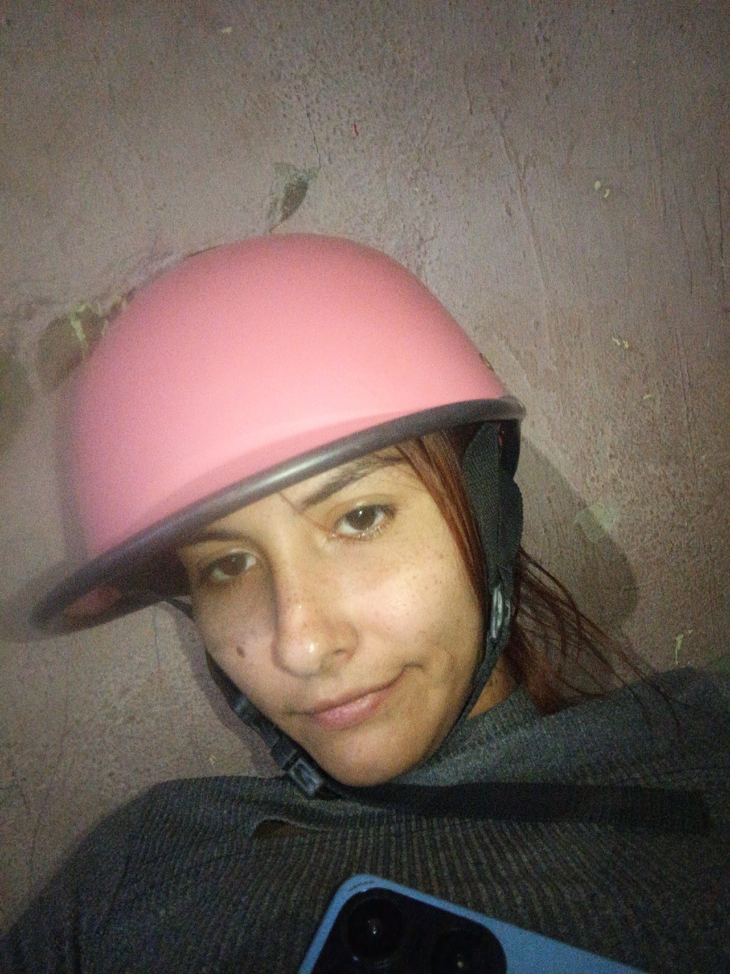 A close-up, selfie-style image shows a young adult female wearing a pink helmet with a gray brim and a black strap. She has brown eyes, freckles, and a slight smile. She is wearing a gray sweater. Part of a blue smartphone is visible at the bottom of the frame. The background is a textured, light gray wall. There is no indication that the picture was taken in the city of Juan Griego, Venezuela.