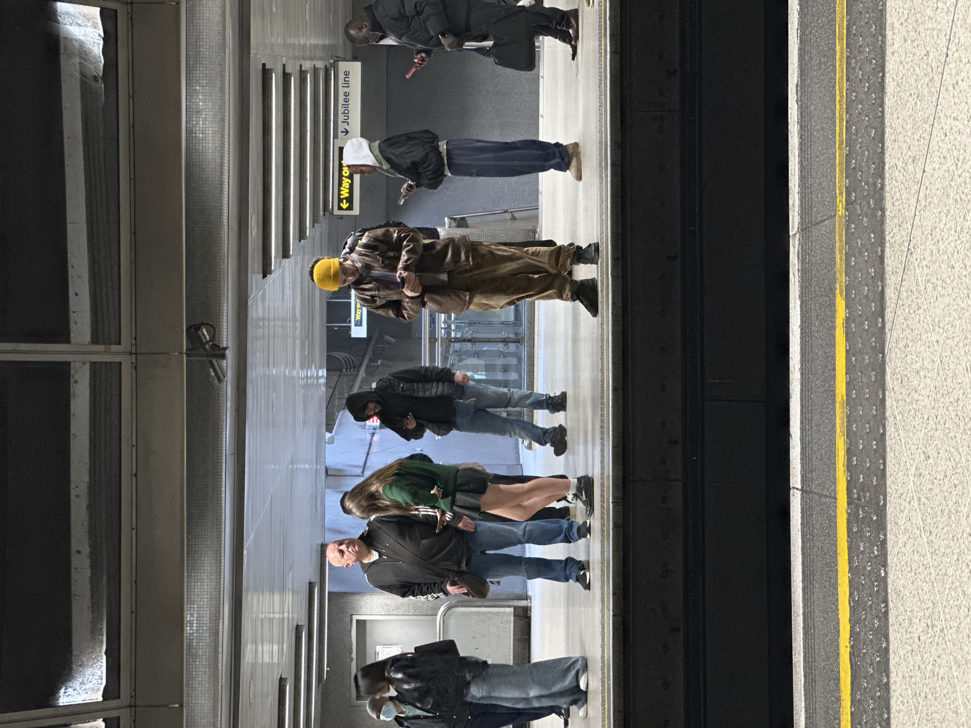 The image depicts a London Underground station platform, specifically indicating the Jubilee Line via an overhead sign displaying "← Way out" and "↓ Jubilee line".
Seven individuals are present on the platform, positioned behind the yellow tactile warning strip. No train is visible.
From left to right:
1. A person in a light blue mask, light jacket, and dark trousers, facing right.
2. A woman in a black leather jacket, dark top, and wide-leg blue jeans, carrying a black shoulder bag, facing right. She appears to be interacting with the person next to her.
3. A bald man in a dark Adidas zip-up hoodie, white t-shirt, and blue jeans, wearing black sneakers, holding a brown shoulder bag. He is looking forward, slightly right.
4. A young woman with long dark hair, wearing a dark green long-sleeved top, a black pleated mini-skirt, white socks, and dark shoes. She is holding a smartphone and looking down at it, facing left.
5. A man wearing a yellow beanie, a brown leather jacket, a grey t-shirt, and brown trousers, with dark sneakers. He is holding a smartphone with both hands and looking down at it, facing forward.
6. A person in a white hoodie, dark jacket, and dark tracksuit bottoms, with light brown shoes. They are holding a smartphone and looking down at it, facing right.
7. A man in a dark puffer jacket and dark trousers, with dark dress shoes. He is holding a red-cased smartphone in his right hand and a black brief-style bag in his left hand, looking down at the phone.
The platform floor is light-colored concrete with a visible yellow safety line and grey tactile paving. The track area is dark. The station ceiling features multiple rows of fluorescent lighting fixtures. White tiled walls are visible in the background, along with a grey metal structure supporting the ceiling. A white door is visible to the left. The scene is illuminated by artificial light, suggesting an indoor or underground setting. The overall atmosphere is one of passive waiting, with most individuals engaged with their mobile devices.