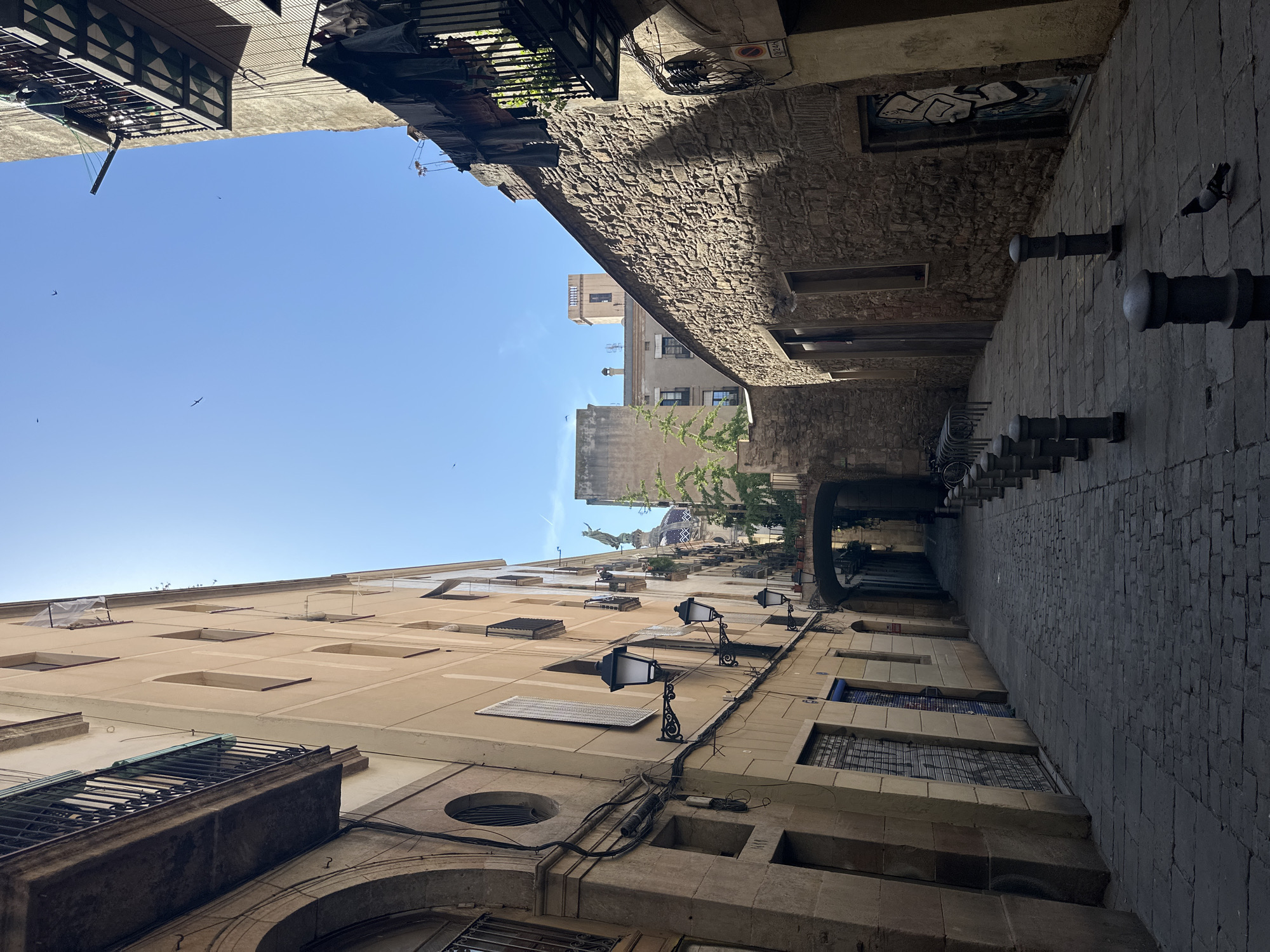 The image captures a narrow, cobblestone street in the Gothic Quarter of Barcelona, Spain, bathed in bright sunlight. The street is flanked by old, tall buildings with beige and stone facades. On the left, a building features arched entrances, balconies with wrought-iron railings, and several windows. Decorative street lamps with ornate black metal brackets are affixed to its side. The right side of the street is dominated by a rough-hewn stone wall with a few small windows and a door covered in graffiti. A wrought-iron balcony with laundry hanging from it extends from an upper floor on the right.
The street narrows as it recedes into the distance, leading under a stone archway. A line of dark, rounded bollards runs down the center of the street, creating a pedestrianized area. In the background, a glimpse of a building with a dome and a statue of an angel is visible, suggesting a church or cathedral. The sky is a clear, bright blue with a few small birds in flight. A single pigeon is present in the foreground on the cobblestone pavement. The overall impression is one of historic charm and urban density.