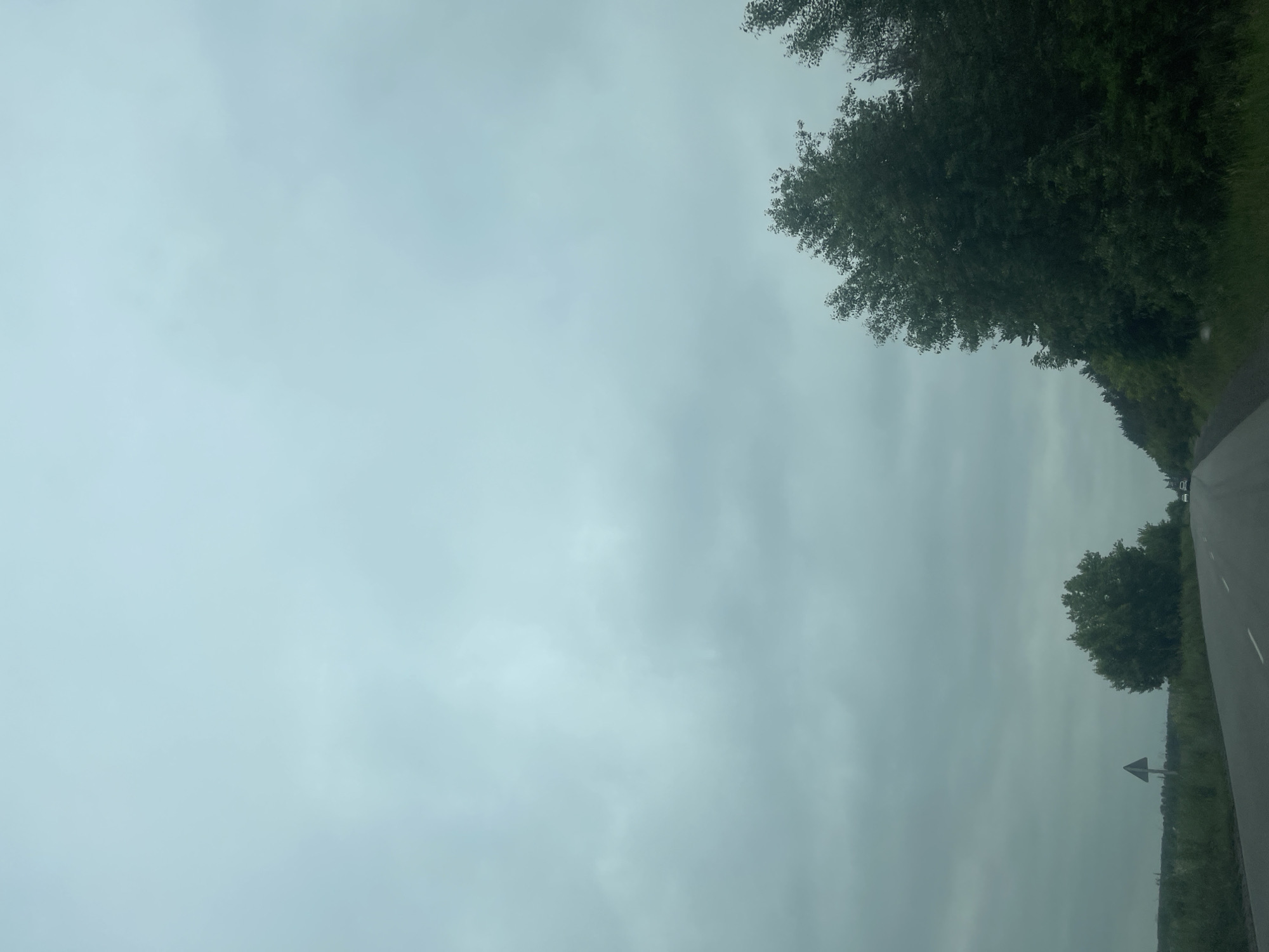 The image depicts a view from a vehicle traveling on a paved road. The sky is overcast with a muted, grayish-blue hue, suggesting a cloudy day. Dense, dark green foliage lines the right side of the road, with trees that appear lush and full. On the left side of the road, there is a grassy verge with a triangular road sign. In the distance, the road stretches towards the horizon, where it is flanked by fields or lower vegetation. A few vehicles are visible on the road in the distance. The scene appears to be a rural or semi-rural area, consistent with a location in or near Копили, Ukraine.