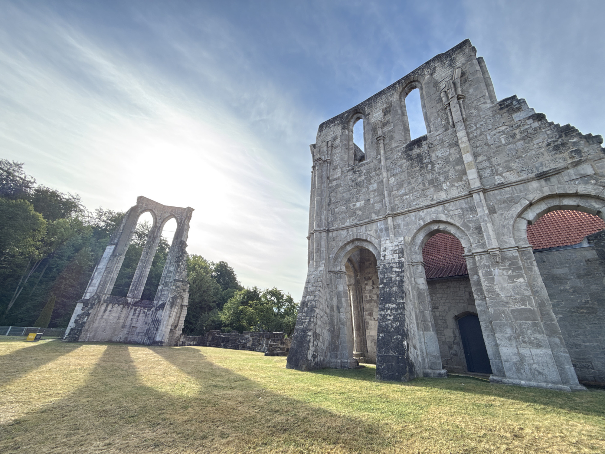 The image shows the ruins of the Walkenried Abbey in Walkenried, Germany, under a bright, cloudy sky. The ruins are made of stone and feature arched windows and doorways. One large section of the ruins dominates the right side of the frame, with a red-tiled roof visible behind some of the arches. To the left, a smaller, more dilapidated section of the ruins stands against a backdrop of trees. The ground in front of the ruins is covered in dry grass, with long shadows cast by the structures, indicating the sun is low in the sky. No people are visible in the image.