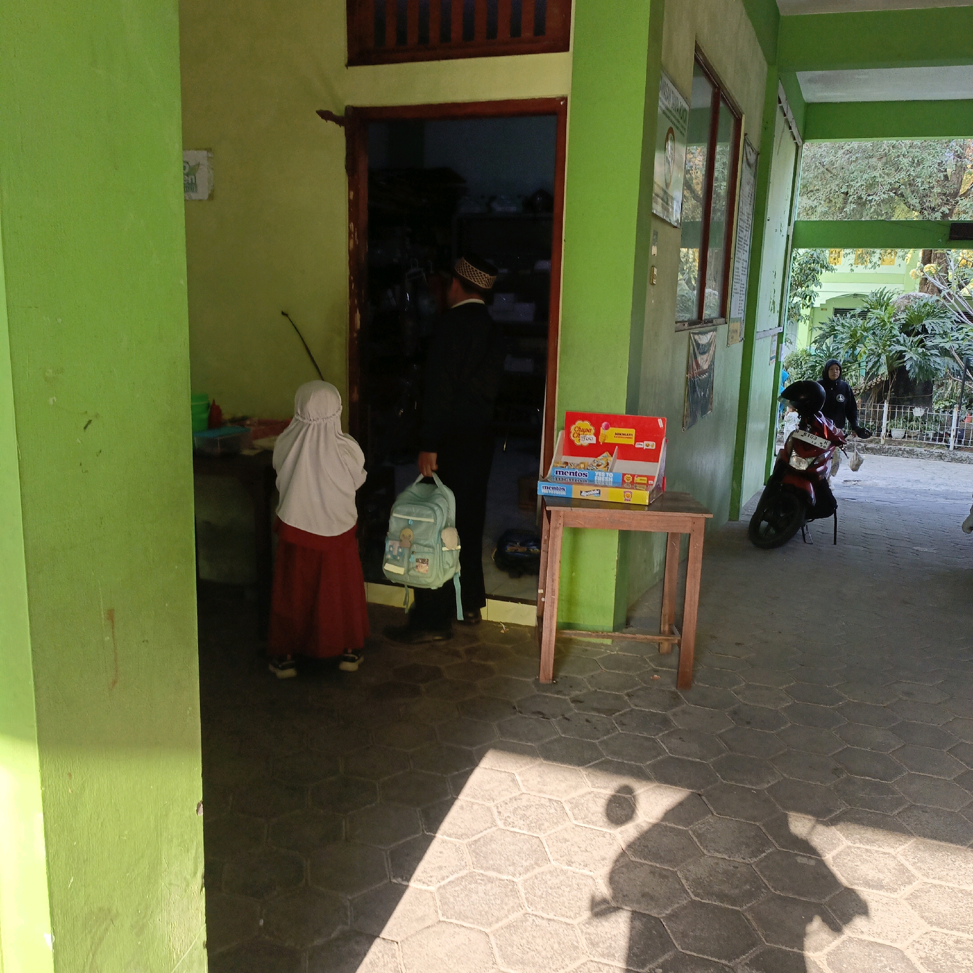In Salatiga, Indonesia, a young student wearing a white hijab and a red skirt stands outside a building with green walls. Adjacent to the student, a person in a black suit and cap is visible through an open doorway. The student carries a light blue backpack adorned with cartoon figures. A wooden table stands to the right of the doorway, holding a display of Mentos candies and a red box with the Chupa Chups logo. Beyond the building, a red motorcycle is parked, and a person in a black hooded jacket is standing nearby. The ground is paved with hexagonal tiles.