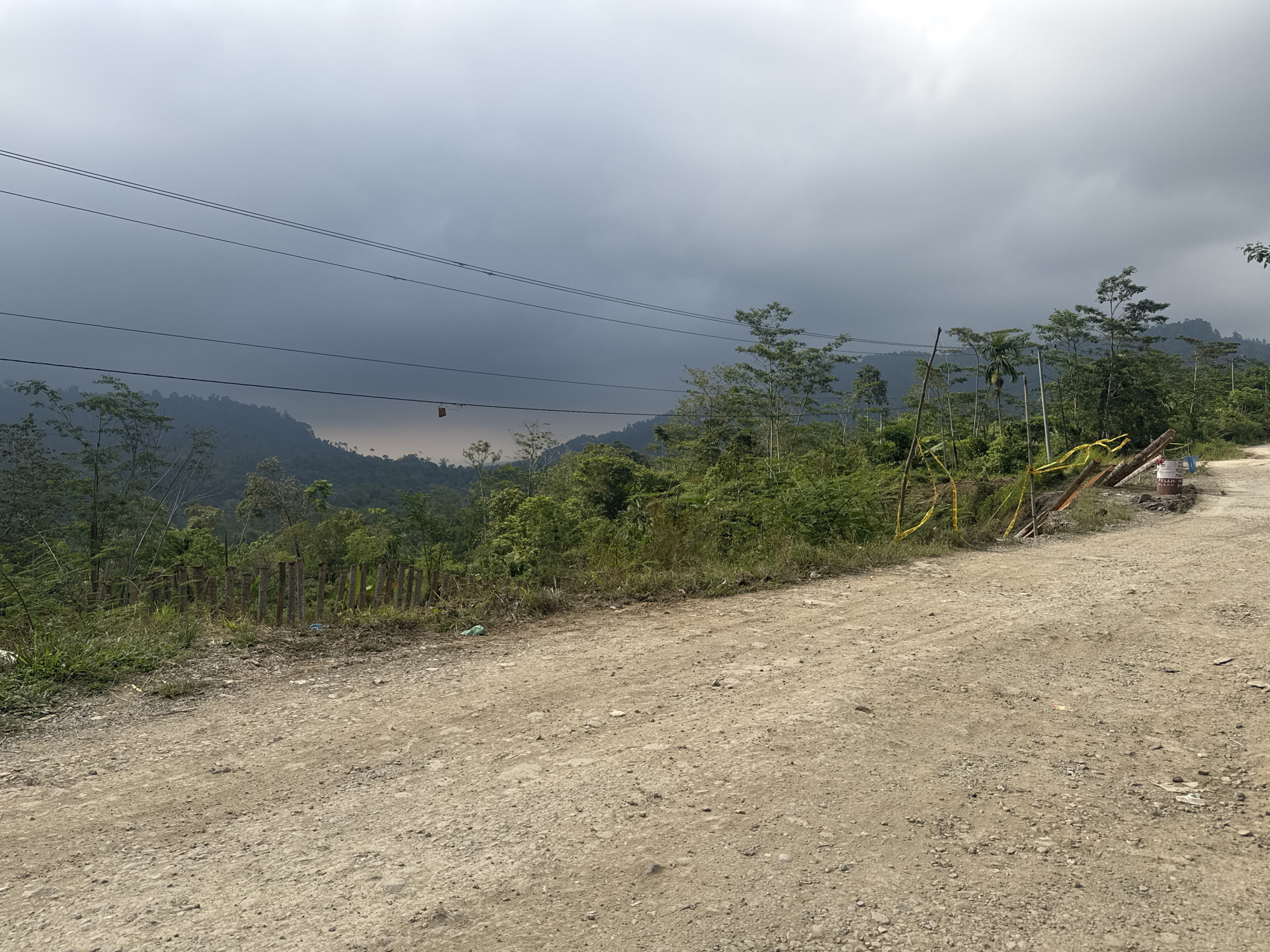 This is a wide, landscape, eye-level shot of a dusty, unpaved road winding through a rural, mountainous area. The sky is overcast and dark, with a heavy layer of clouds that suggest impending rain. In the foreground, the road is made of dirt and gravel, with some scattered rocks and debris. To the left of the road, there is a steep embankment covered in green grass and vegetation. A makeshift fence made of wooden posts is visible along the edge of the embankment. Further back, a dense forest covers the rolling hills and mountains that recede into the distance. Power lines are strung across the upper portion of the frame, running horizontally above the trees. A small, red object, possibly a hanging ornament or marker, is attached to one of the power lines. Yellow caution tape is strung between wooden posts on the right side of the road, indicating ongoing work or a hazard. A white and red cylindrical object, possibly a paint can or a sealant container, is also visible on the right side of the road, near the caution tape. The overall impression is one of a remote, undeveloped area experiencing overcast weather conditions.
