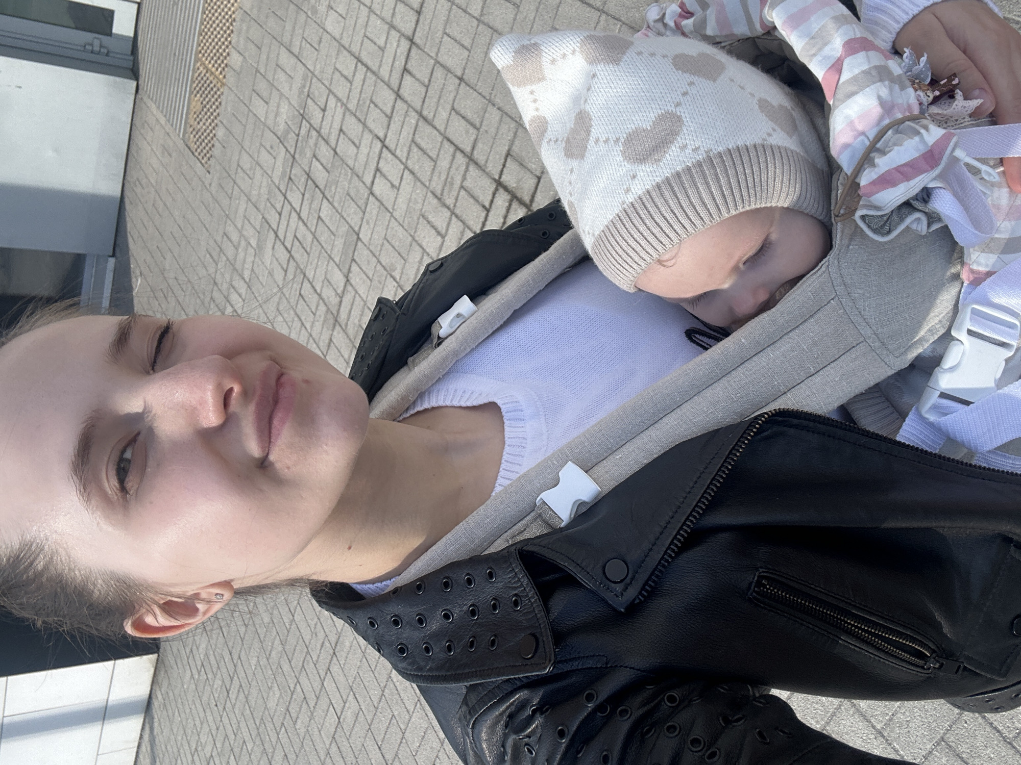 The image displays an outdoor, eye-level selfie-style photograph taken on a sunny day in Khimki, Russia. A young adult female is centered, holding an infant in a soft-structured baby carrier against her chest. She has fair skin, hair pulled back, and a slight smile with her right eye partially closed. She wears a black leather jacket with numerous circular perforations and studs, over a white knitted sweater. A small stud earring is visible in her right earlobe. The infant, appearing to be asleep with eyes closed, wears a beige and white patterned knitted bonnet. A striped pink, white, and beige fabric partially covers the infant. The baby carrier is light beige with white plastic buckles. The ground is paved with light grey rectangular pavers arranged in a herringbone pattern. In the background, a modern, light-colored building with distinct shadows is visible to the left, and a building wall with a door frame and a metallic ground grate is visible to the right.