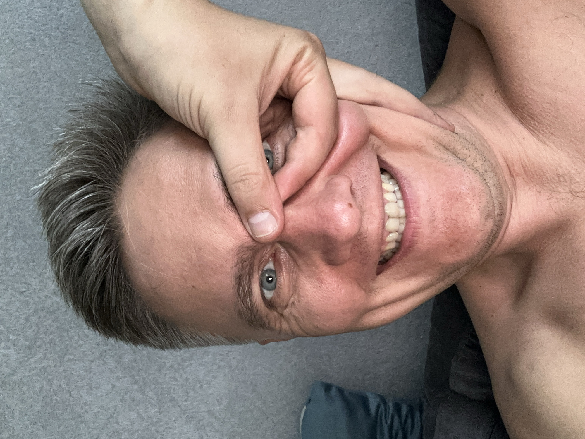 A man lies on a gray surface. His head is positioned towards the left side of the frame, and he is looking at the viewer with a smile. His right hand is pinching his nose. He has fair skin, graying hair, and blue eyes. He appears to be topless. The background seems to be a gray, possibly fabric, surface.
The information on location is not applicable, as the image does not provide sufficient details to identify the specific location.