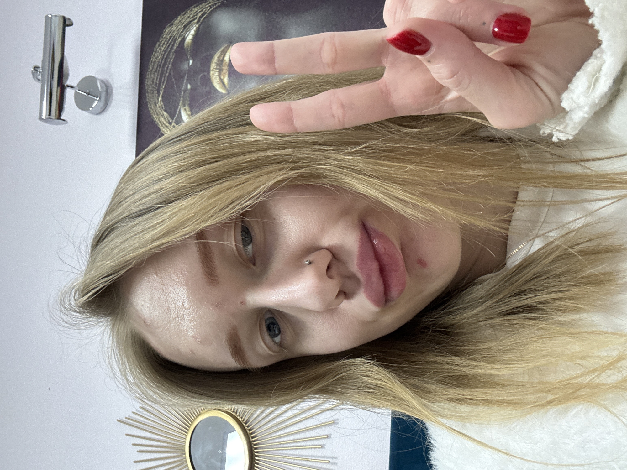 A selfie of a young woman with blonde hair and blue eyes, showing a peace sign with red nail polish. She has a nose piercing and a mark on her lip. The background includes a wall light fixture, a decorative golden sunburst mirror, and a piece of art depicting a face. The location is impossible to determine as nothing in the image indicates the city of Rostov-on-Don.