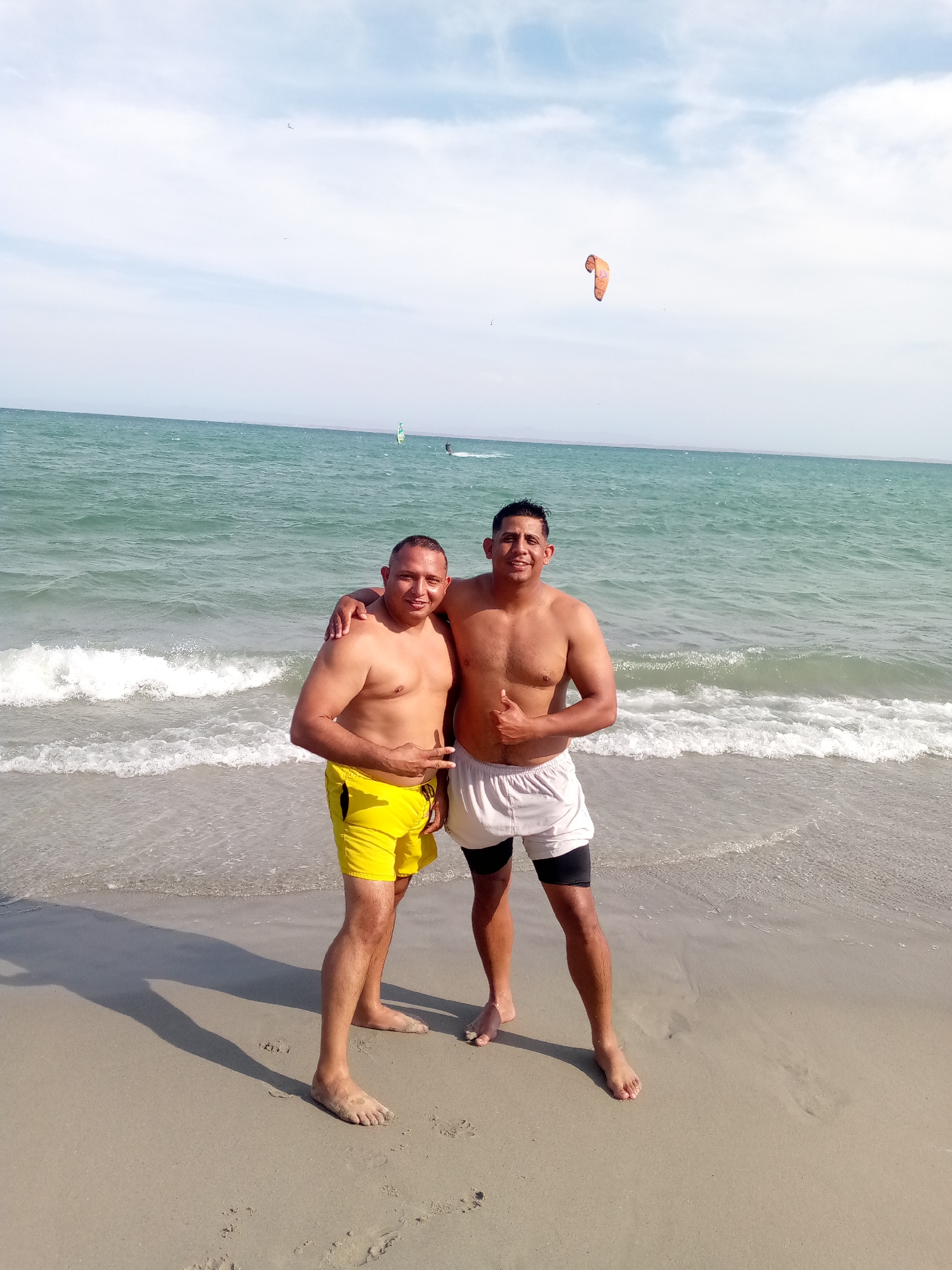 Two men are standing on a sandy beach near the ocean. The man on the left is wearing yellow swim trunks and making a "peace" sign with his fingers. The man on the right is wearing white swim trunks over black compression shorts and giving a "thumbs up" gesture with his right hand. They are standing side-by-side with their arms around each other. Behind them, there is a body of water with small waves, and further in the distance, there is a person windsurfing. Above them, there is a kite in the sky. The image appears to be taken in El Yaque, Venezuela.