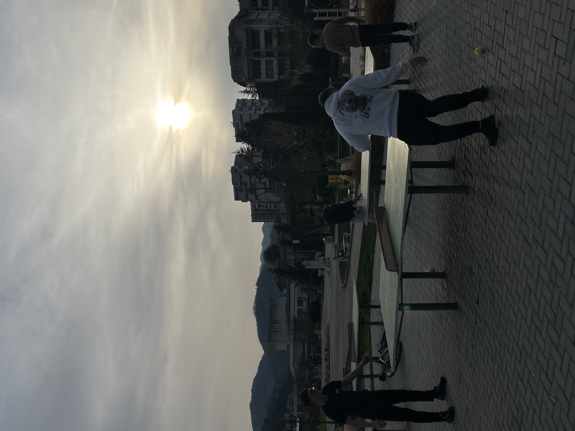 The image shows a scene in the city of Zlatograd, Bulgaria. Four people are visible in an outdoor setting. One person is wearing a white hoodie and black pants and appears to be near a table. Another person is standing nearby wearing casual clothing. A third person is standing in the middle ground and a fourth person is at the bottom of the frame. The setting includes buildings, trees, a brick paved area, and what appears to be mountains or hills in the background. The sky is overcast, with the sun partially obscured. There is a ball on the paved area.