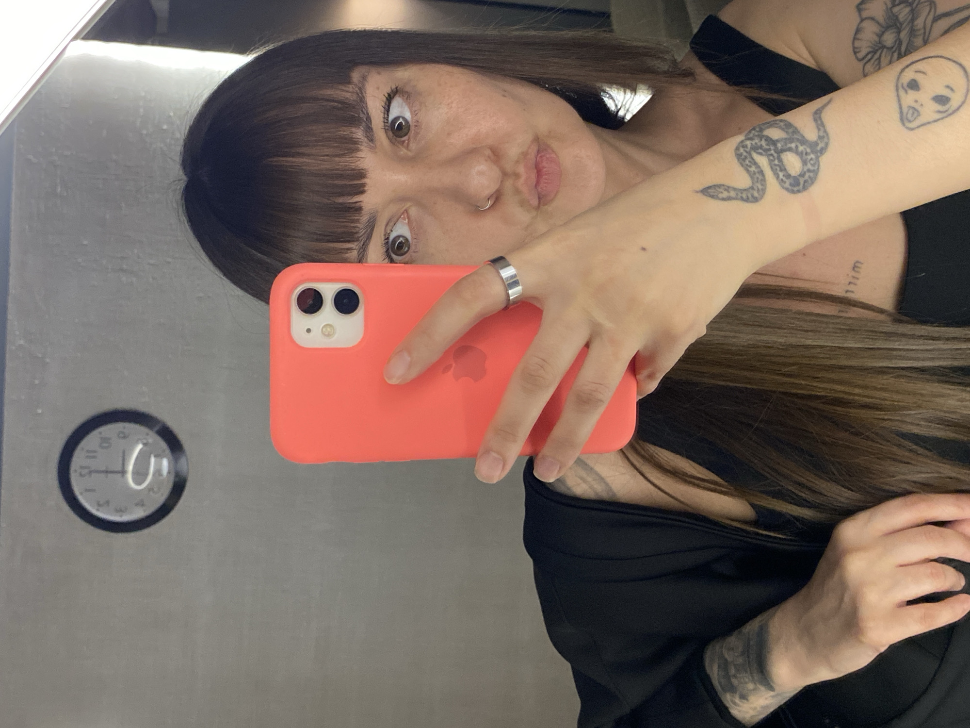 A woman with dark hair and bangs takes a selfie in front of a mirrored wall. She holds a coral-colored iPhone in her right hand, with a silver ring on her ring finger. Her left arm is raised, displaying tattoos including a snake and an alien. A clock on the wall reflects 12:50. The woman's expression is surprised or playful, with wide eyes and pursed lips. The background is a plain, gray wall. This scene is likely in Votkinsk, Russia based on the prompt.