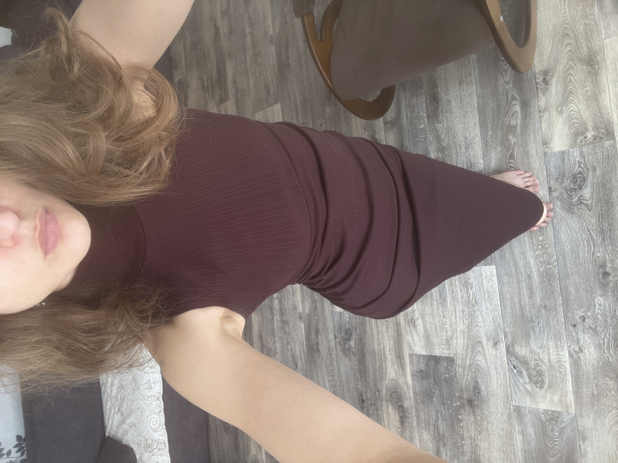 A woman stands in a room with wood-laminate flooring. She is wearing a dark maroon, sleeveless, turtleneck dress that is fitted to her body. Her hair is brown and wavy, and it falls around her shoulders. Her feet are bare, and she is standing on the floor. There is a dark-colored sofa with a white patterned throw blanket in the background, and a brown wooden chair is visible to her right. The image does not provide enough information to determine the specific location in the city of Sharkan, Russia.