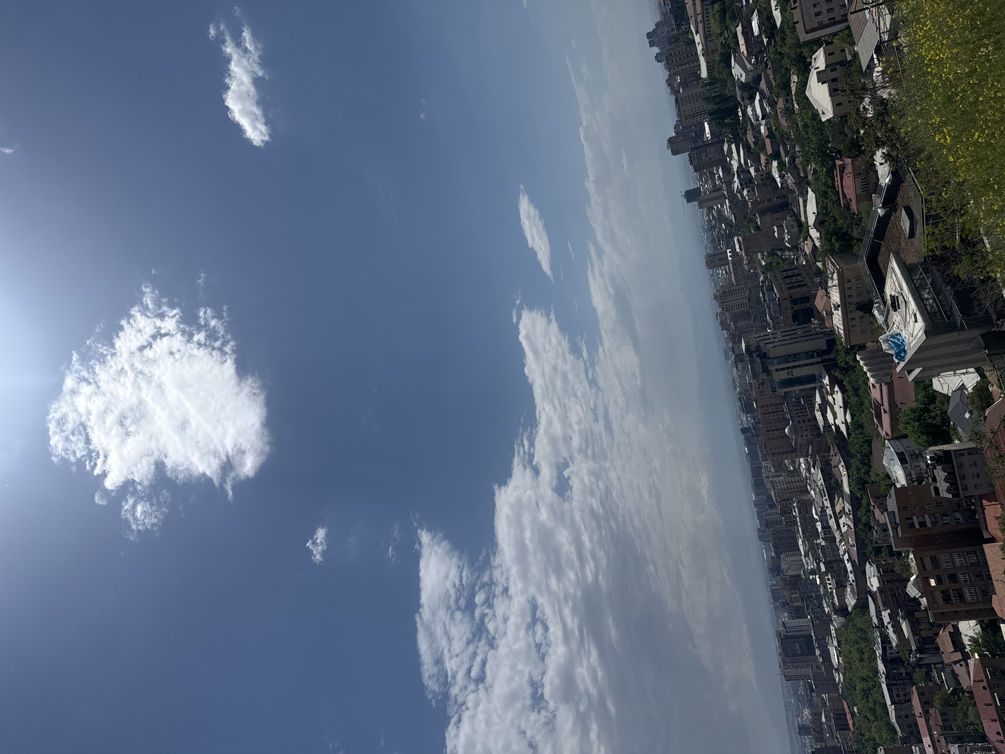 The image shows a view of the city of Yerevan, Armenia, from an elevated position. The sky occupies a significant portion of the image, featuring a blue expanse with scattered white clouds of varying sizes and shapes. The sun is visible in the upper left corner of the image, suggesting a bright, clear day.
The lower part of the image reveals a densely populated urban landscape. Buildings of different sizes and architectural styles are tightly packed together, interspersed with trees. The overall impression is of a vibrant, bustling city.