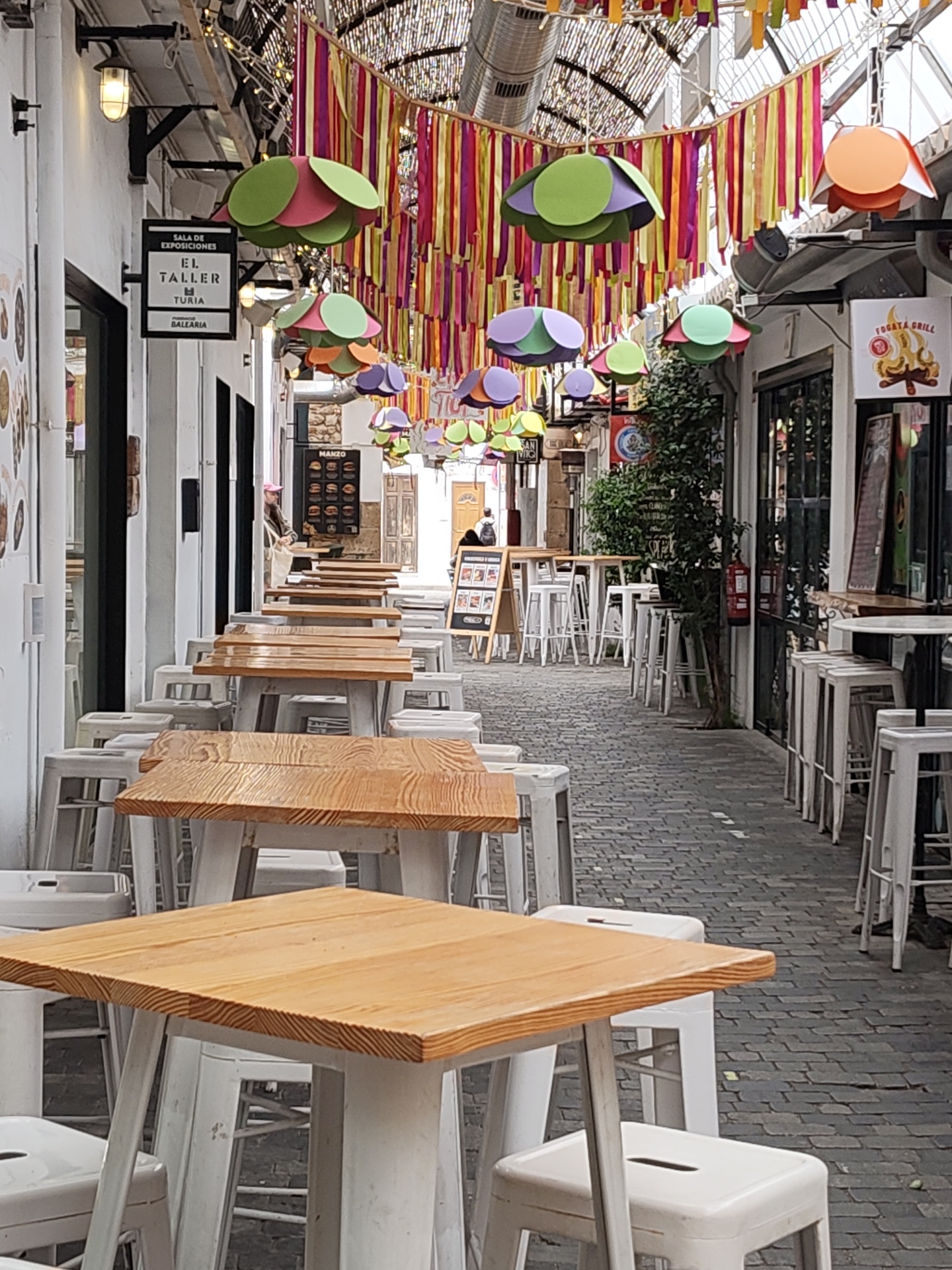The image depicts a narrow pedestrian street in Dénia, Spain, lined with restaurants or cafes. The street is paved with gray bricks. Several rows of tables and white stools extend along the length of the street. The tables have wooden tops. Decorative elements, including colorful hanging ribbons and paper-like flower decorations, are suspended overhead. The buildings on either side are white with black-framed doors and windows. Signs are visible on some of the buildings, including one for "EL TALLER TURIA BALEARIA" and another for "FOGATA GRILL". People are seen in the background, and some are seated. A small tree and a fire extinguisher are located along the right side of the street. The lighting appears to be a combination of natural daylight and artificial lighting.